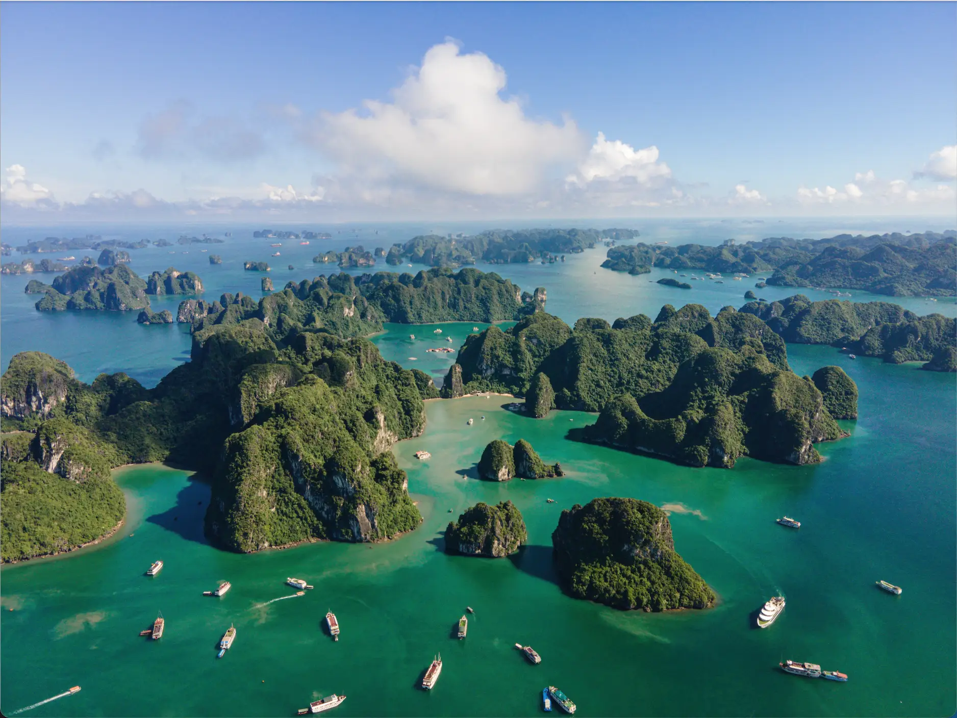 An aerial view of tall mountains on islands, surrounded by emerald-green water and boats.