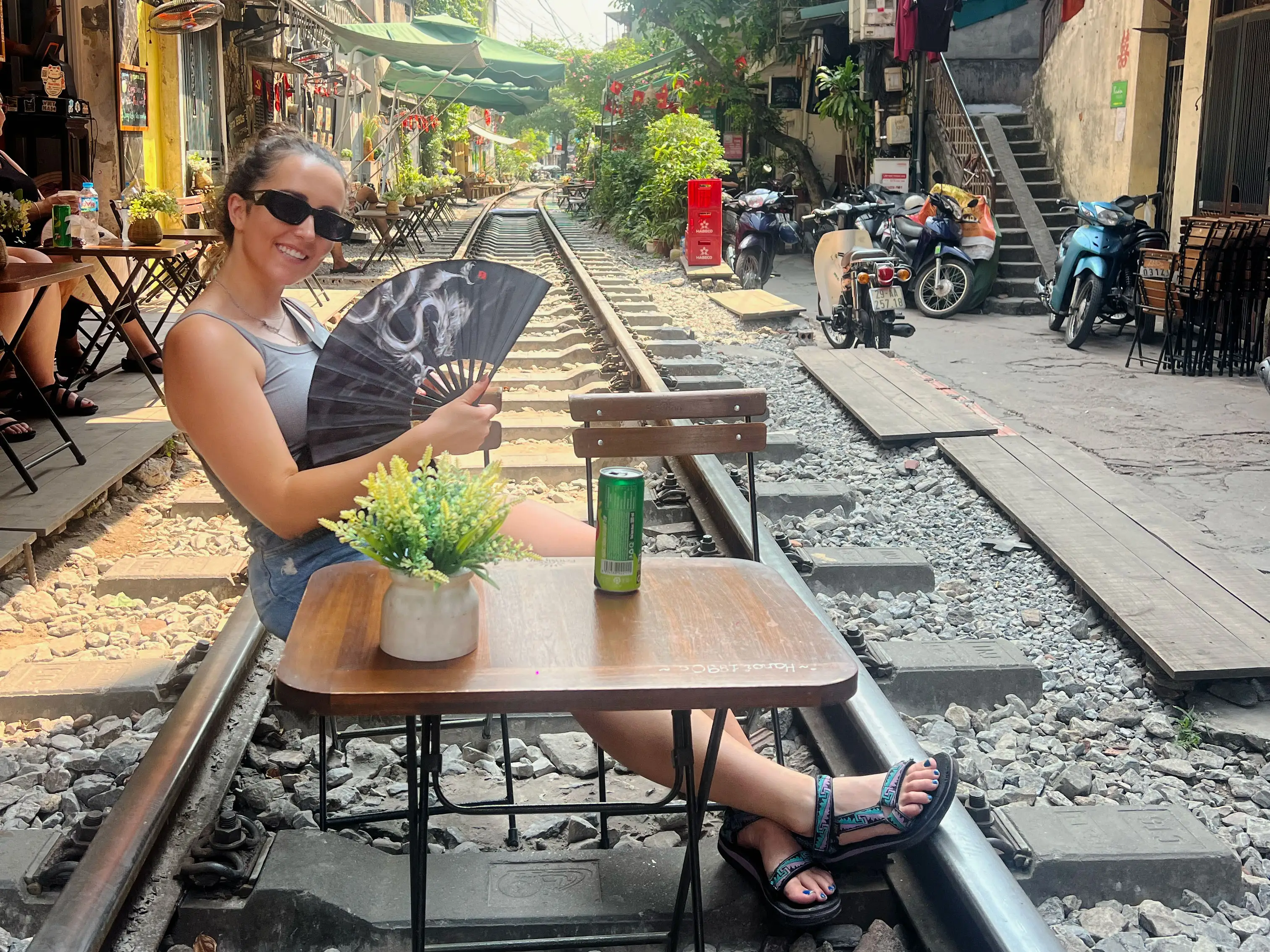 A woman, wearing a tank top, shorts, and sandals, fans herself while sitting at a table on a railway track.