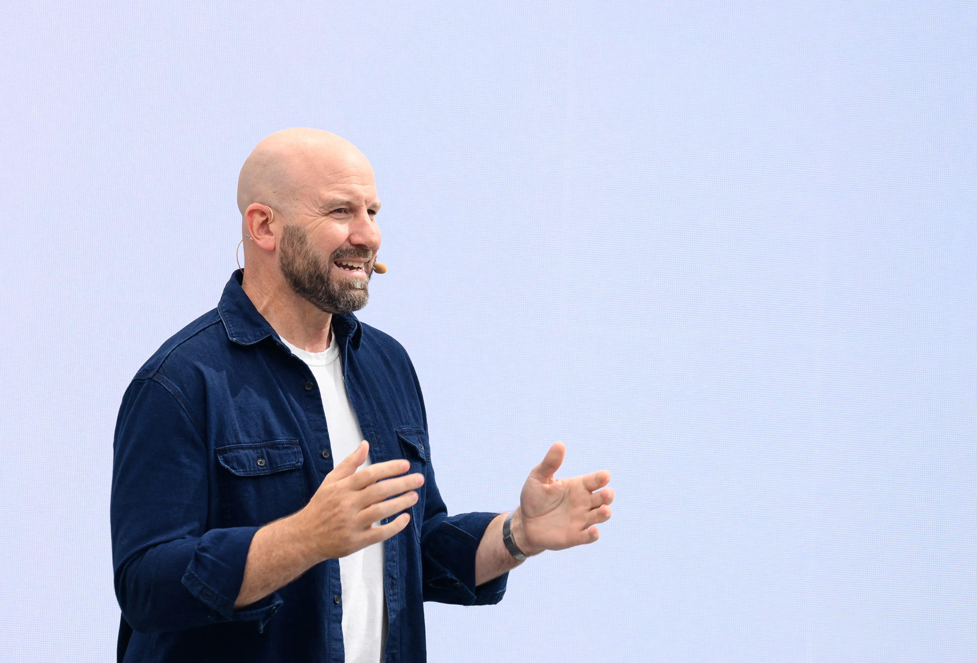 Andrew Bosworth, Meta CTO, is mid speech with his hands held aloft in front of him, as if emphasizing a point. He is standing in front of a light blue background and wearing a dark denim overshirt, which is open over a white undershirt. He is wearing a microphone on his face.