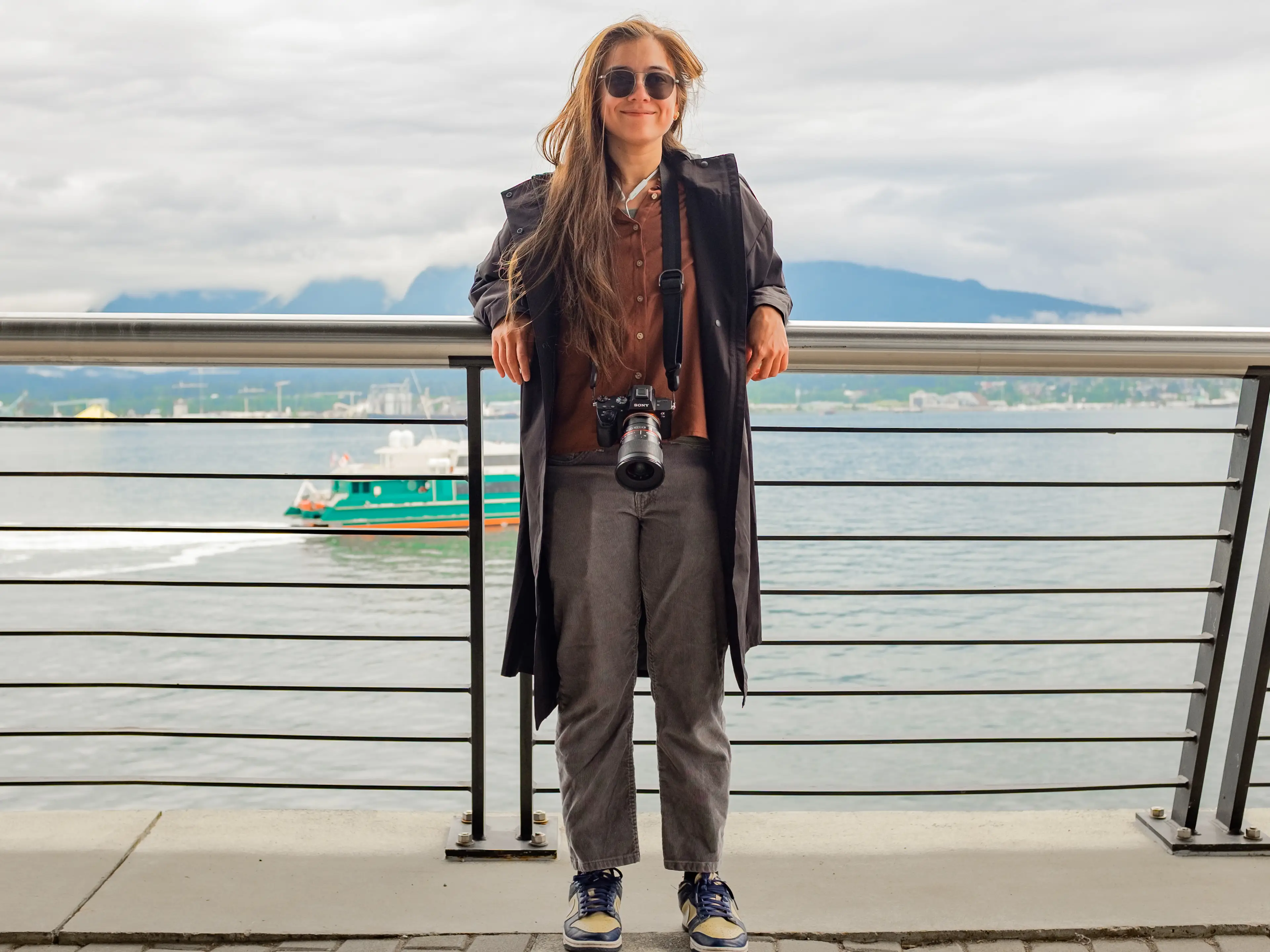 The author leans against a railing in front of the ocean with mountains behind on a cloudy day in Vancouver