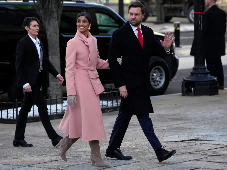 Usha Vance holding onto JD Vance's arm as the walk together on the street. She's wearing a light-pink coat and scarf with tan gloves and boots. He's wearing a black coat and a red tie.