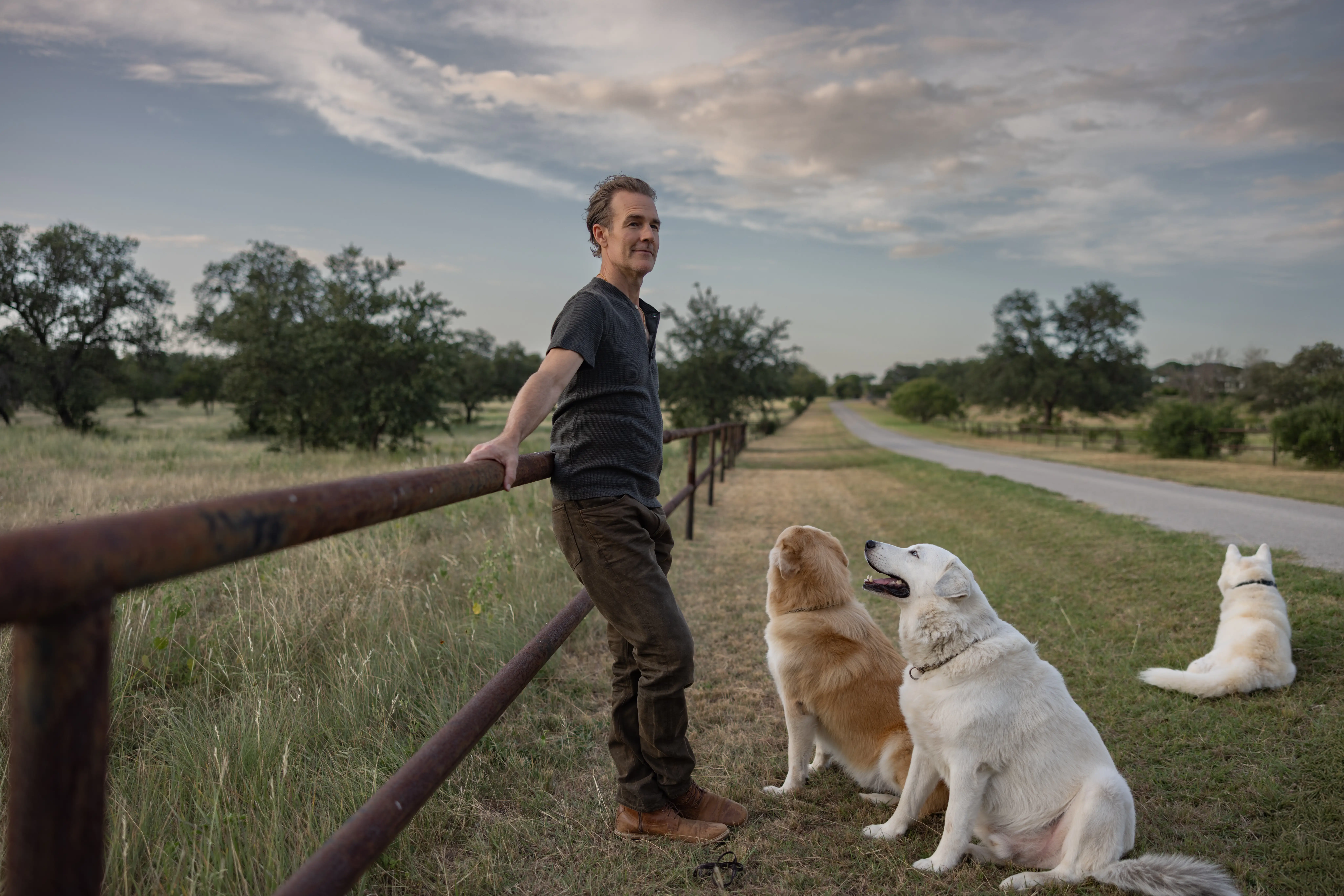 James Van Der Beek leaning against a fence near an open road with three dogs