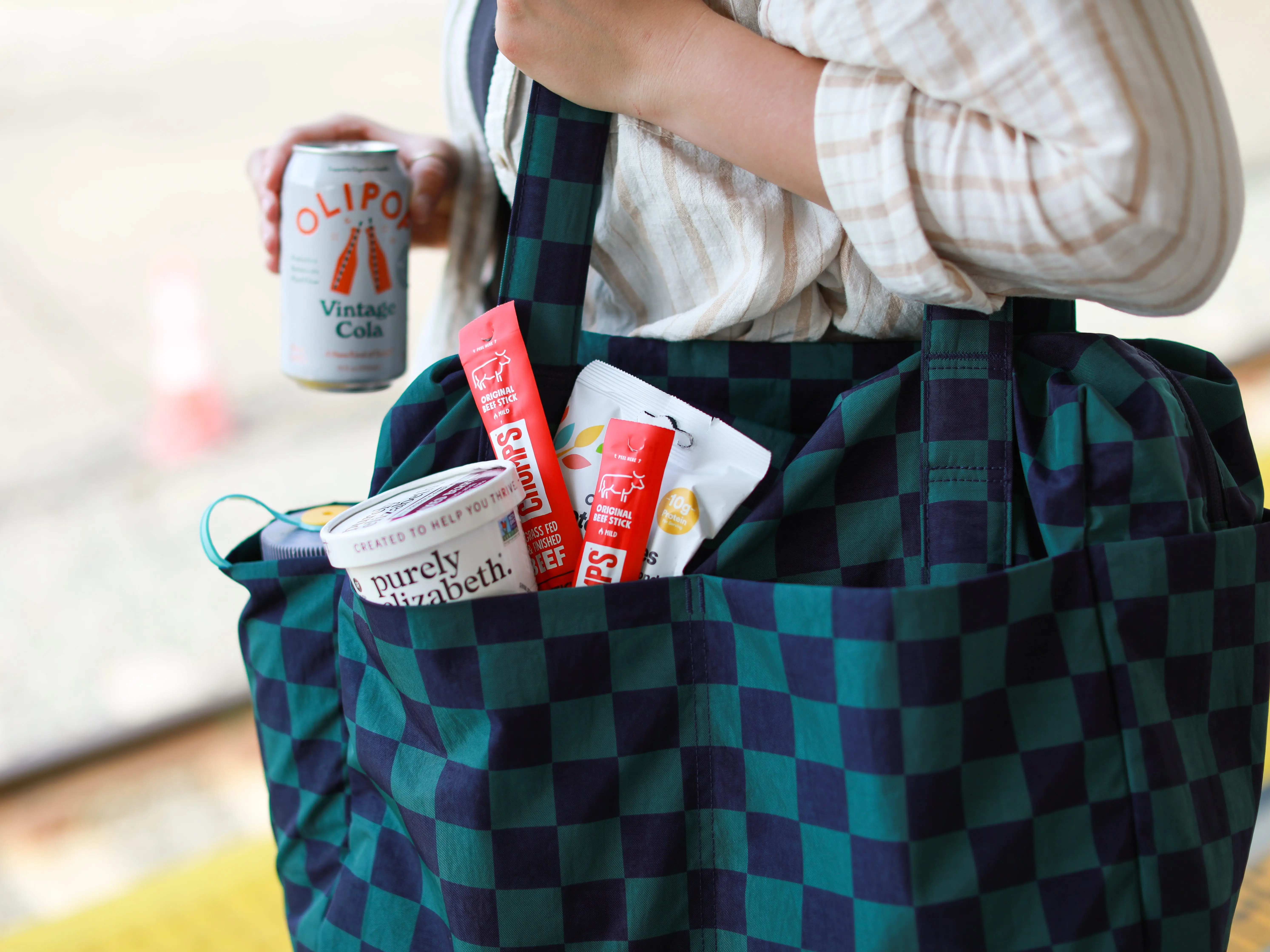 Kylie holds a blue checkered bag with pockets filled with snacks, and a can of Olipop.