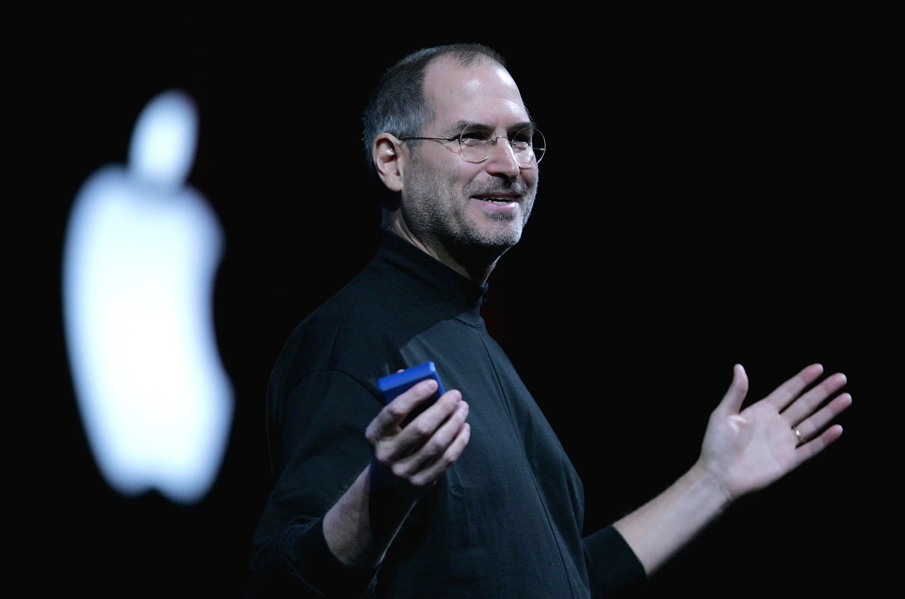 Steve Jobs in a black turtleneck holding an iPhone.