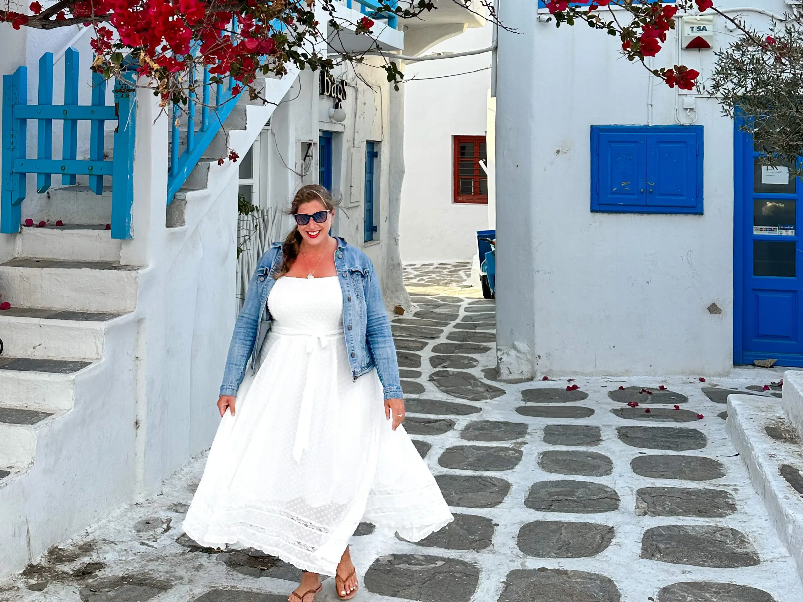 Hannah in a long dress standing on a white stone street, surrounded by  white buildings with blue doors and a red flowering tree overhead.