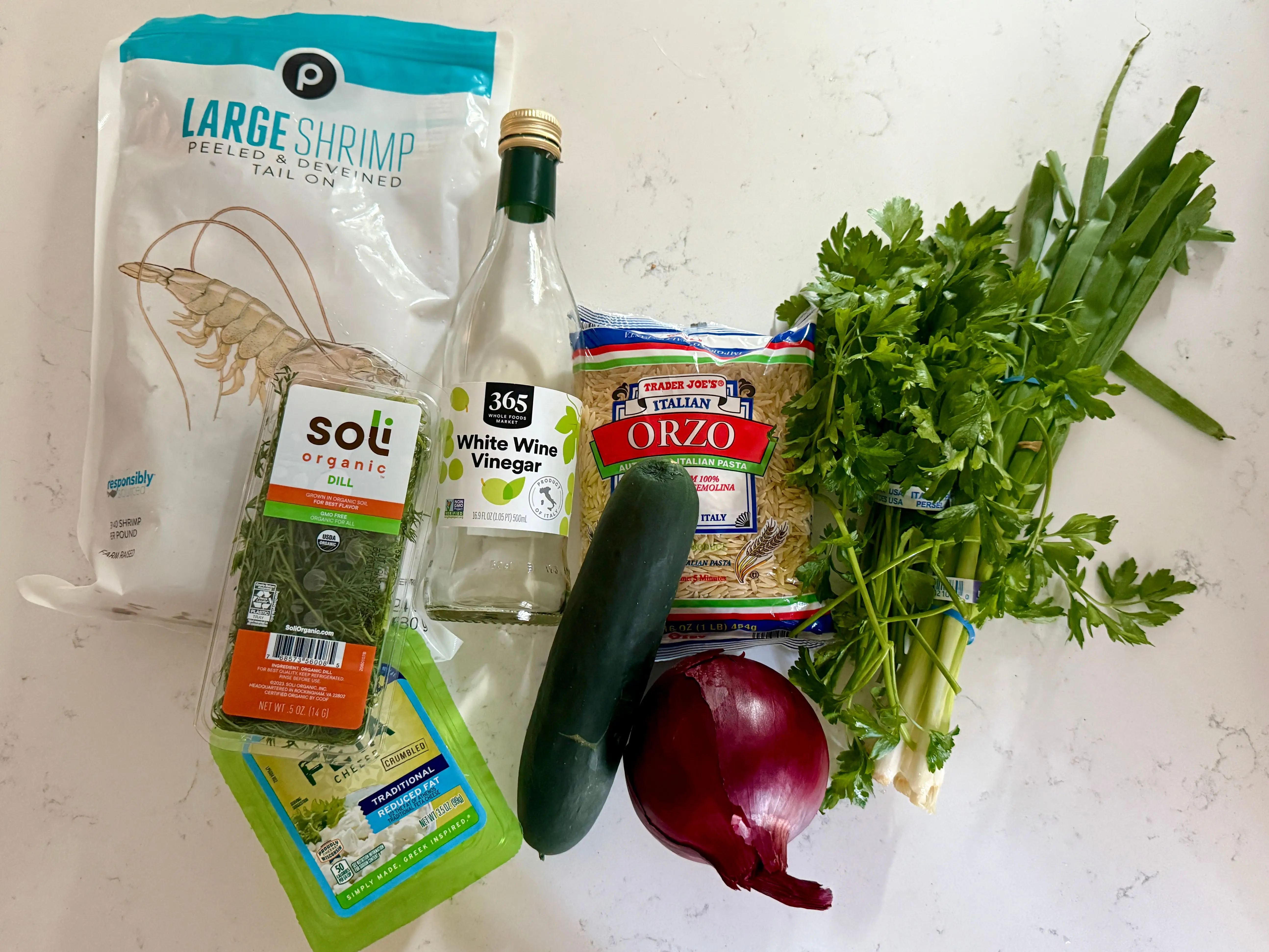A bag of shrimp, a container of dill, a block of feta, a bottle of white wine vinegar, a cucumber, a red onion, a bag of orzo, a bunch of parsley, and a bundle of scallions.