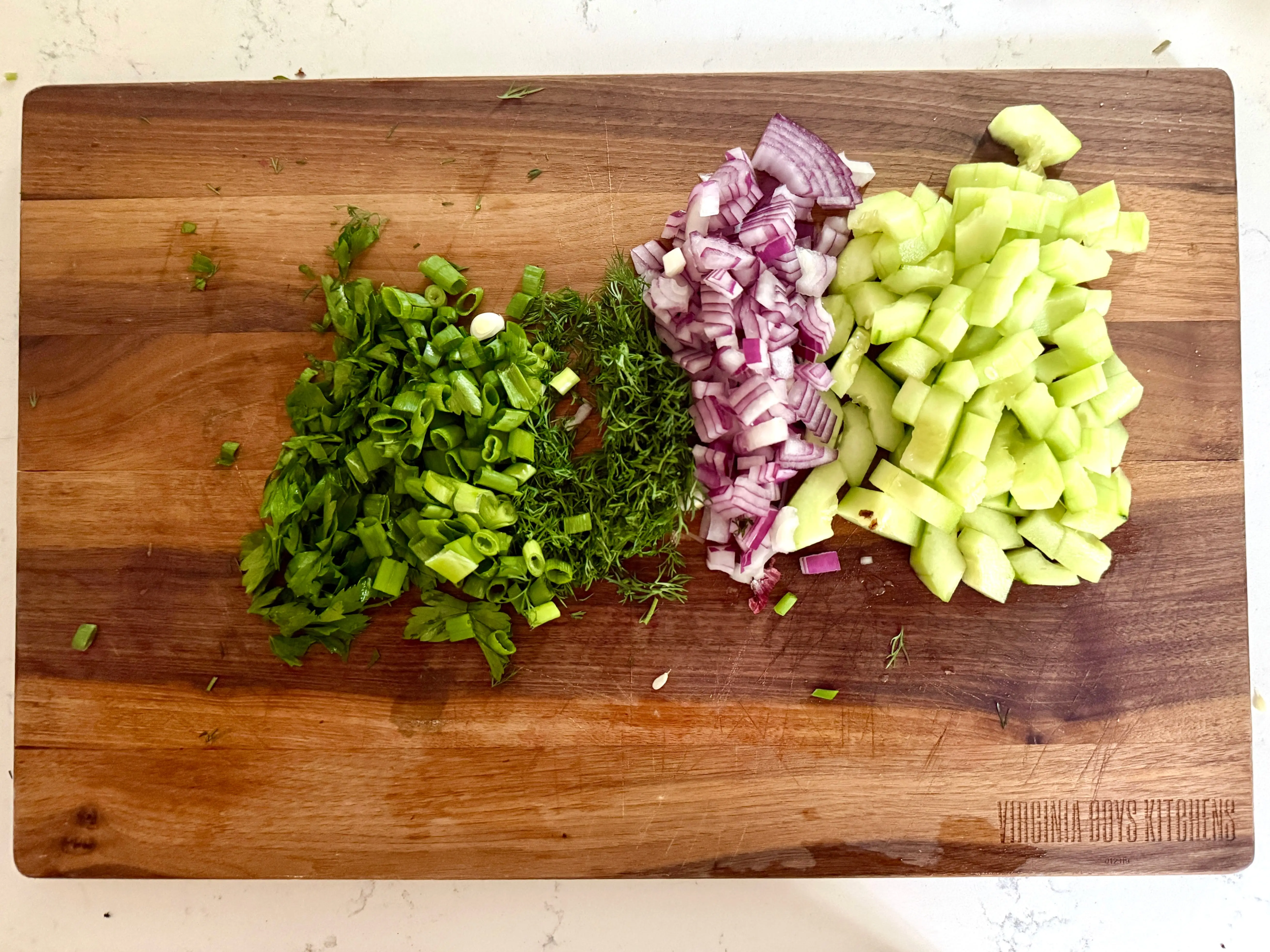 Chopped parsley, scallion, dill, red onion, and cucumber on a wooden cutting board.