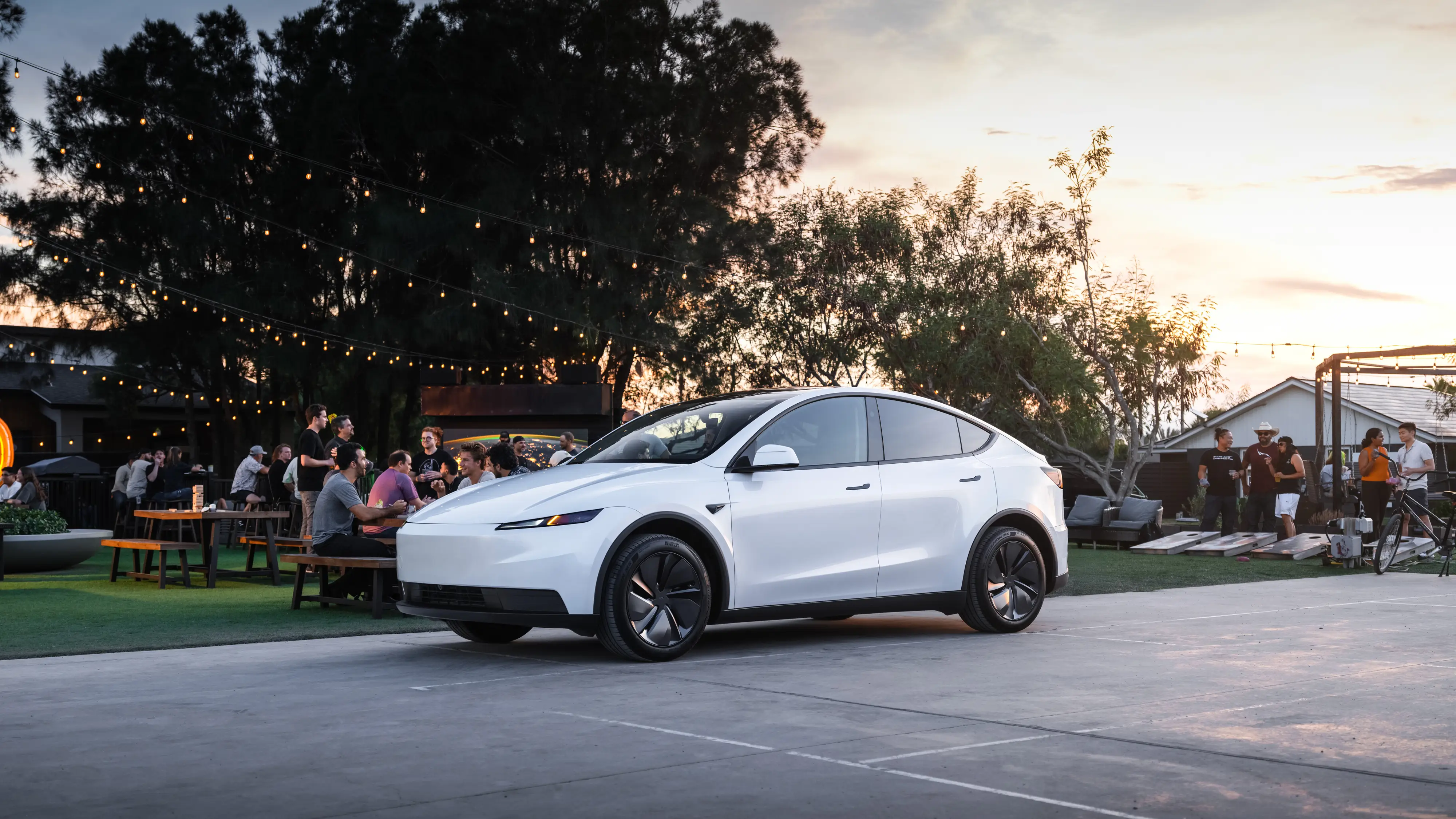 A white Tesla Model Y Standard parked on a gray slab of cement. Behind the car, groups of people are partying with picnic benches and cornhole boards.