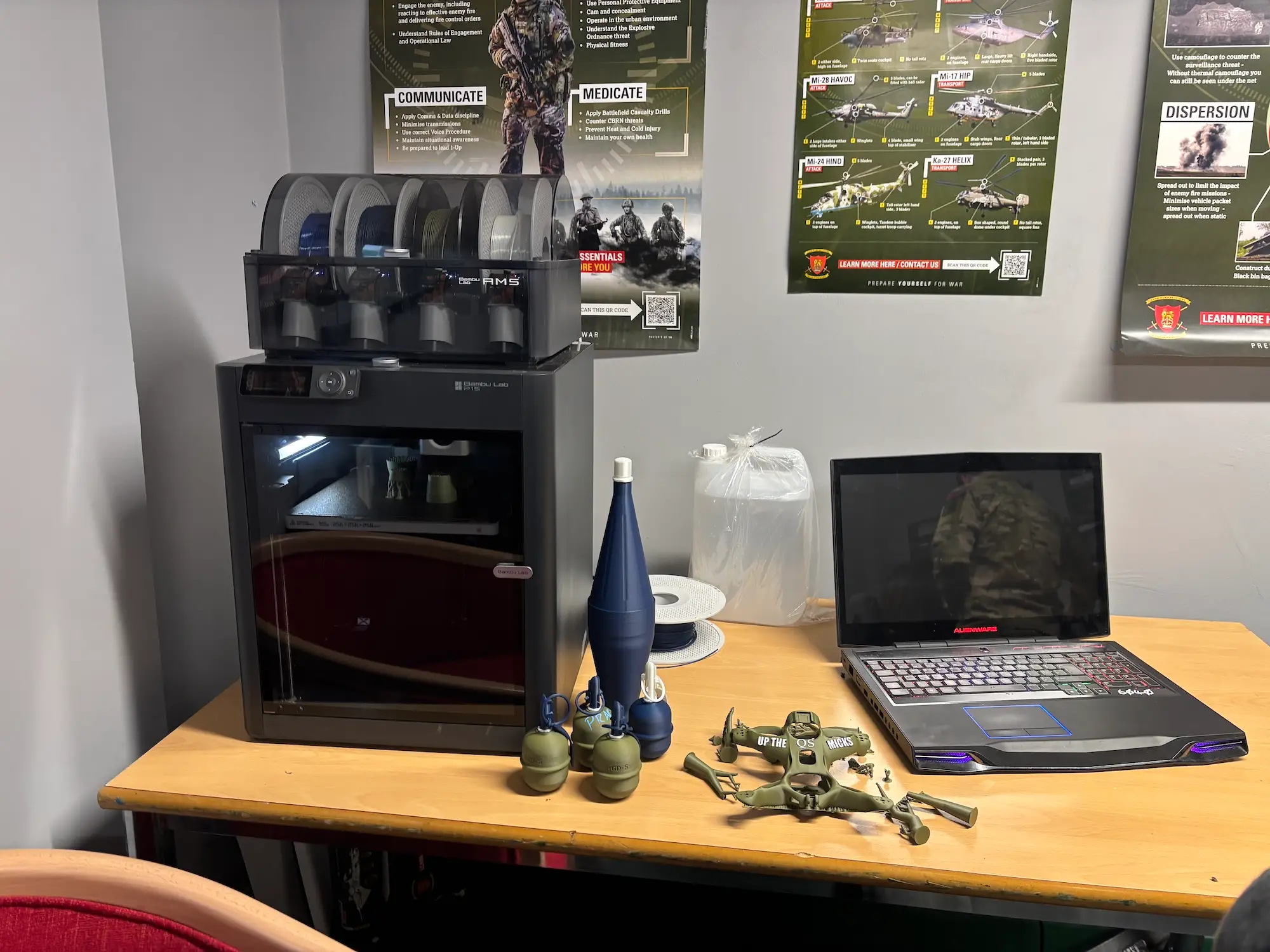 A 3D printer on a table beside a laptop and 3D-printed bomblet shapes with posters on a grey wall that have information about staying alive and Russian weaponry types