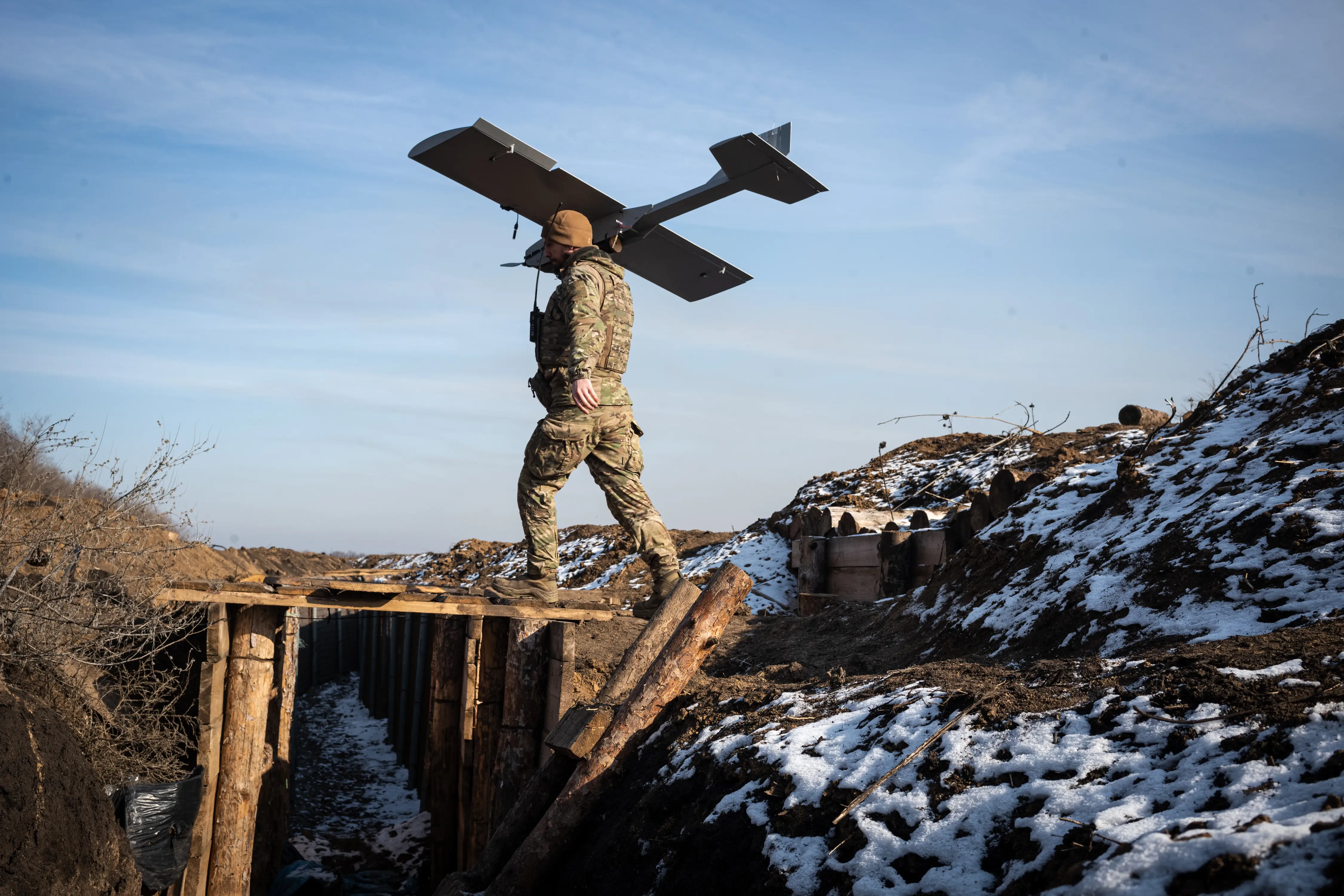 A man in camouflage gear holds a large grey drone on his shoulder walks across a wooden bridge over a trench with a blue sky behind him and snowy ground around him