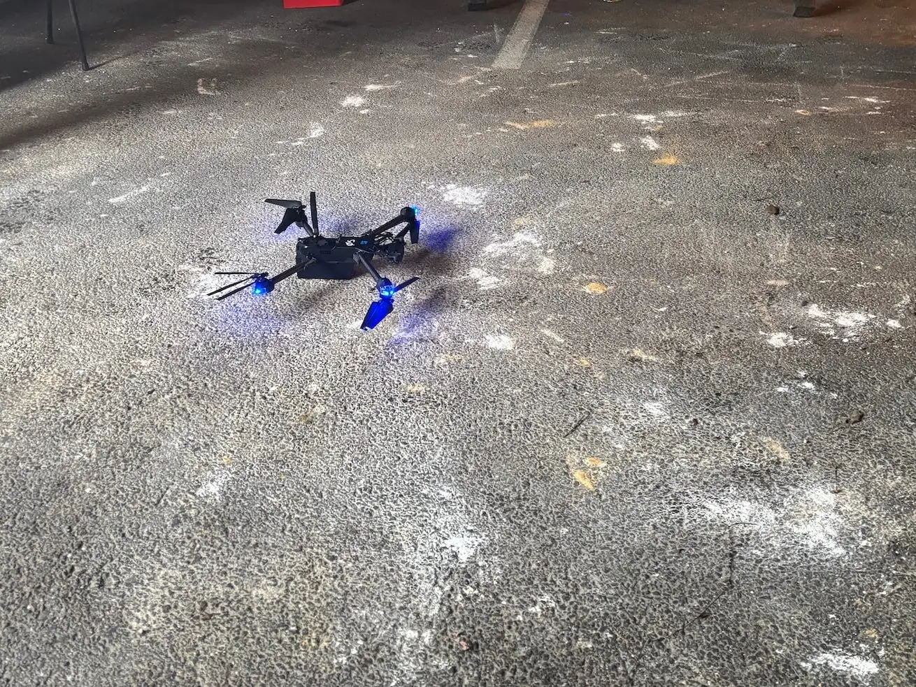 A black small drone with blue lights sits on concrete inside a shed-like room