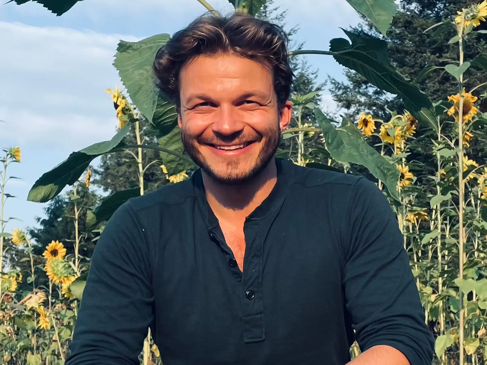 A smiling man sits among tall sunflowers, facing the camera on a sunny day.
