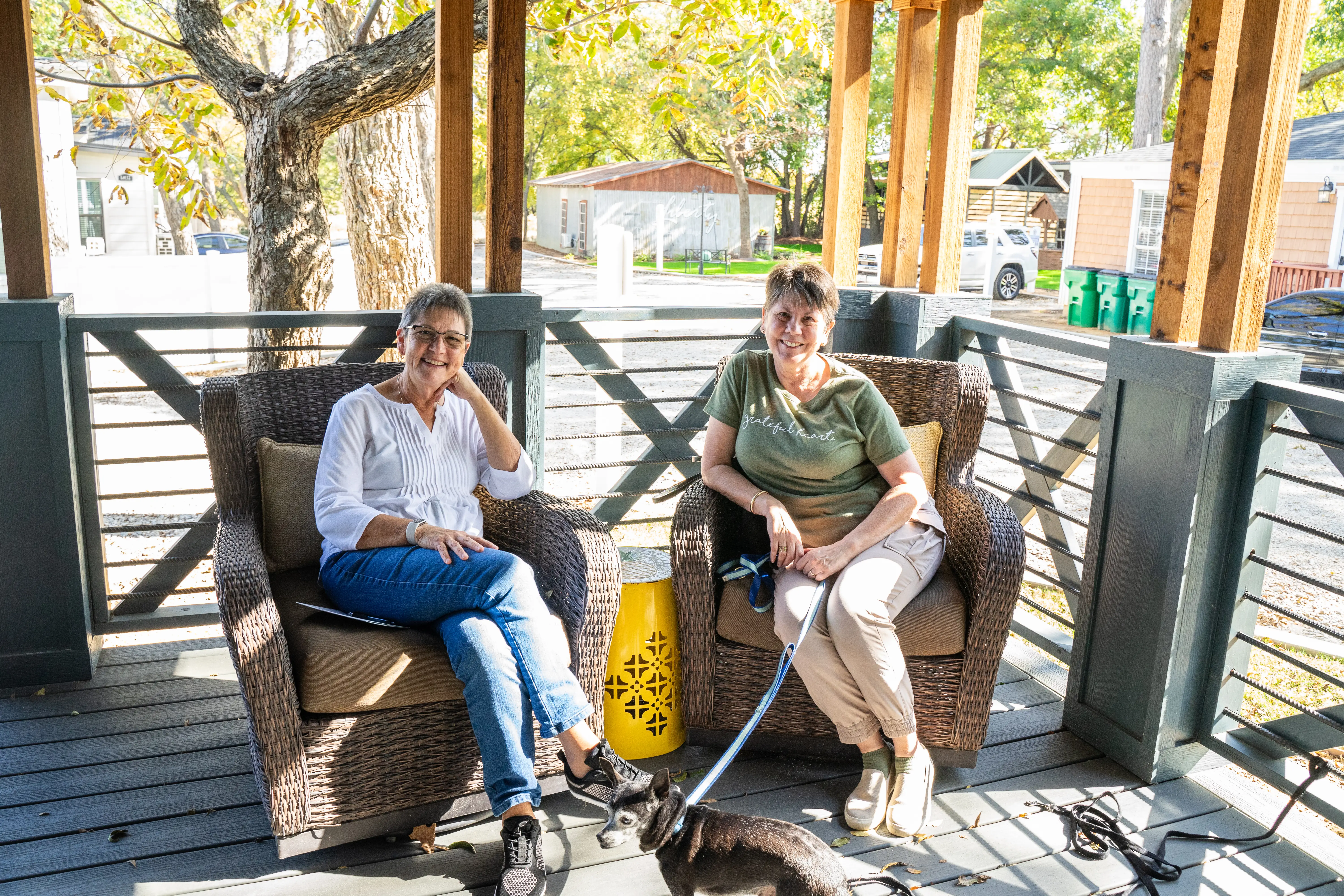 Two women sit on the porch, smiling; one holds a dog on a leash.