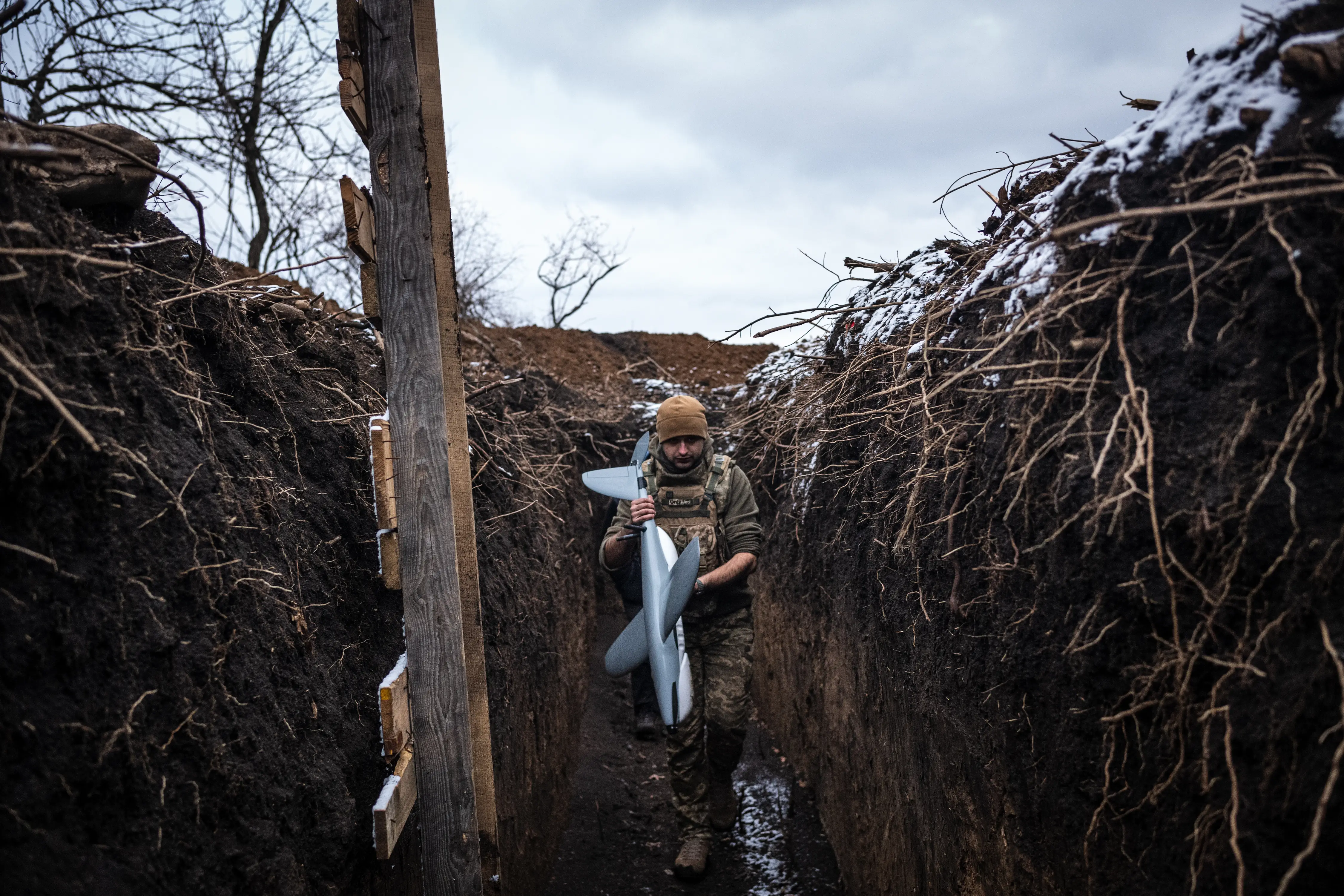 A man crouches in a muddy trench holding a grey drone