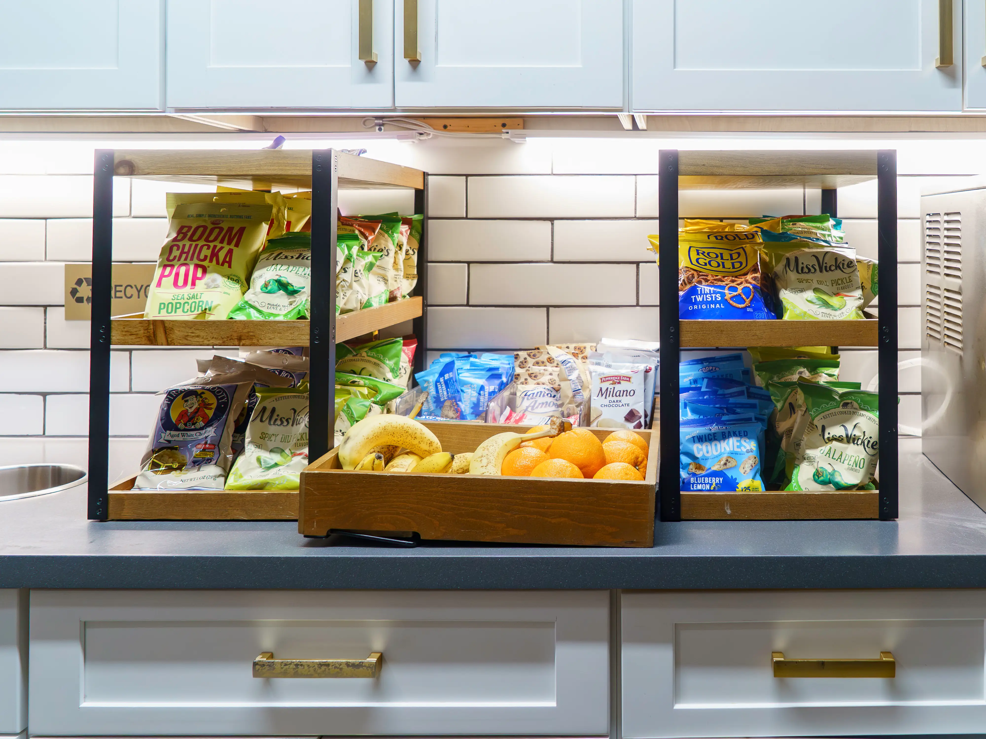 A white kitchen counter has exposed wooden shelves on it stocked with fruit and snacks
