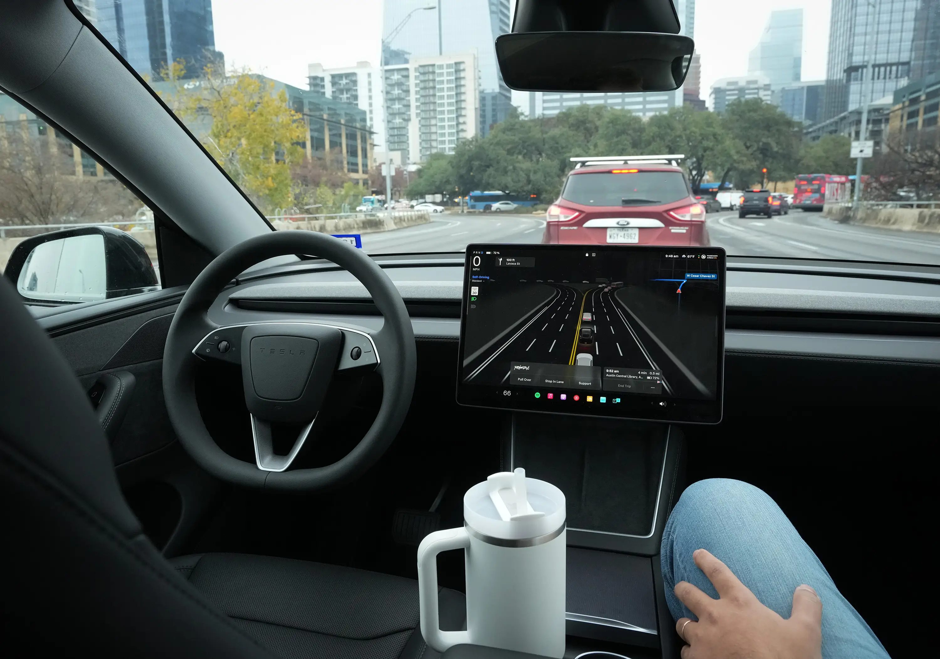 A person in light blue jeans sits in the front passenger seat inside a self-driving Tesla.