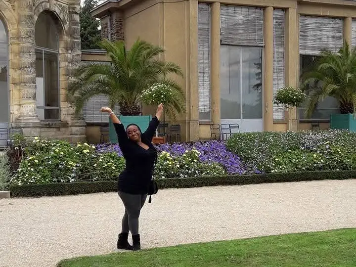 Woman with hands in air smiling on stone path with flowers, building behid her