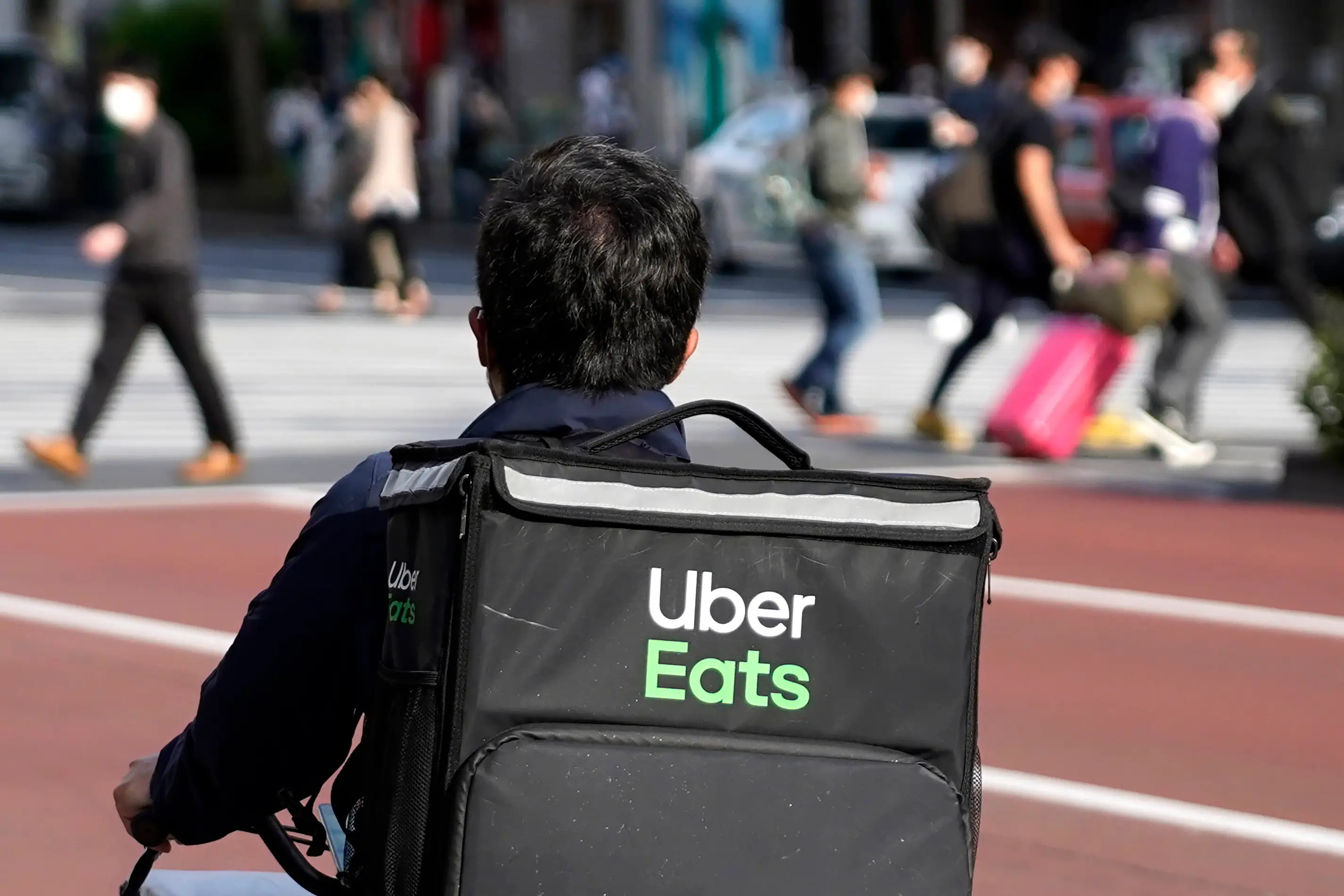 A bike courier wears a black Uber Eats backpack as he approaches an intersection where pedestrians are crossing the street.