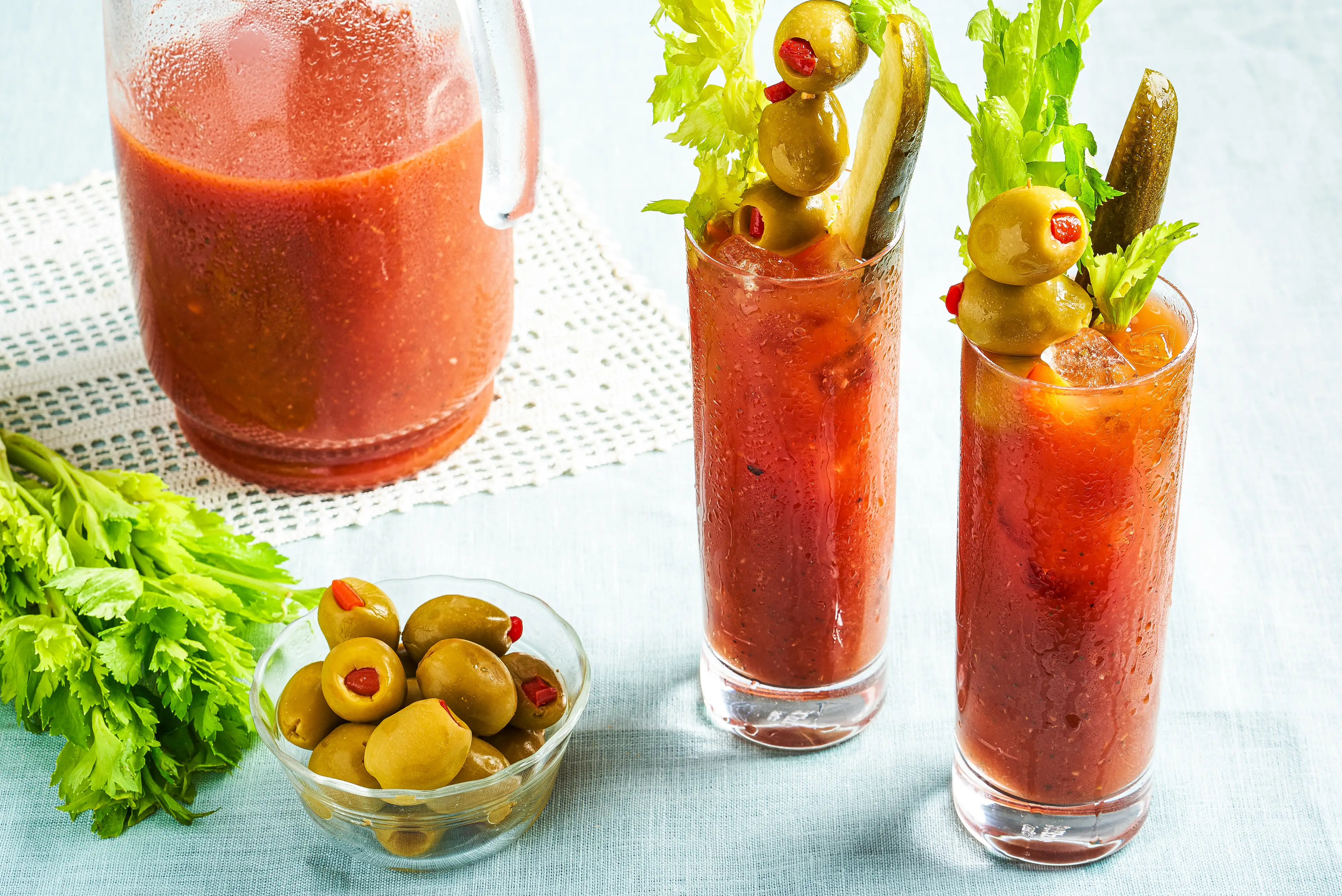 A pitcher and two glasses of Bloody Mary cocktails with olives and pickles.