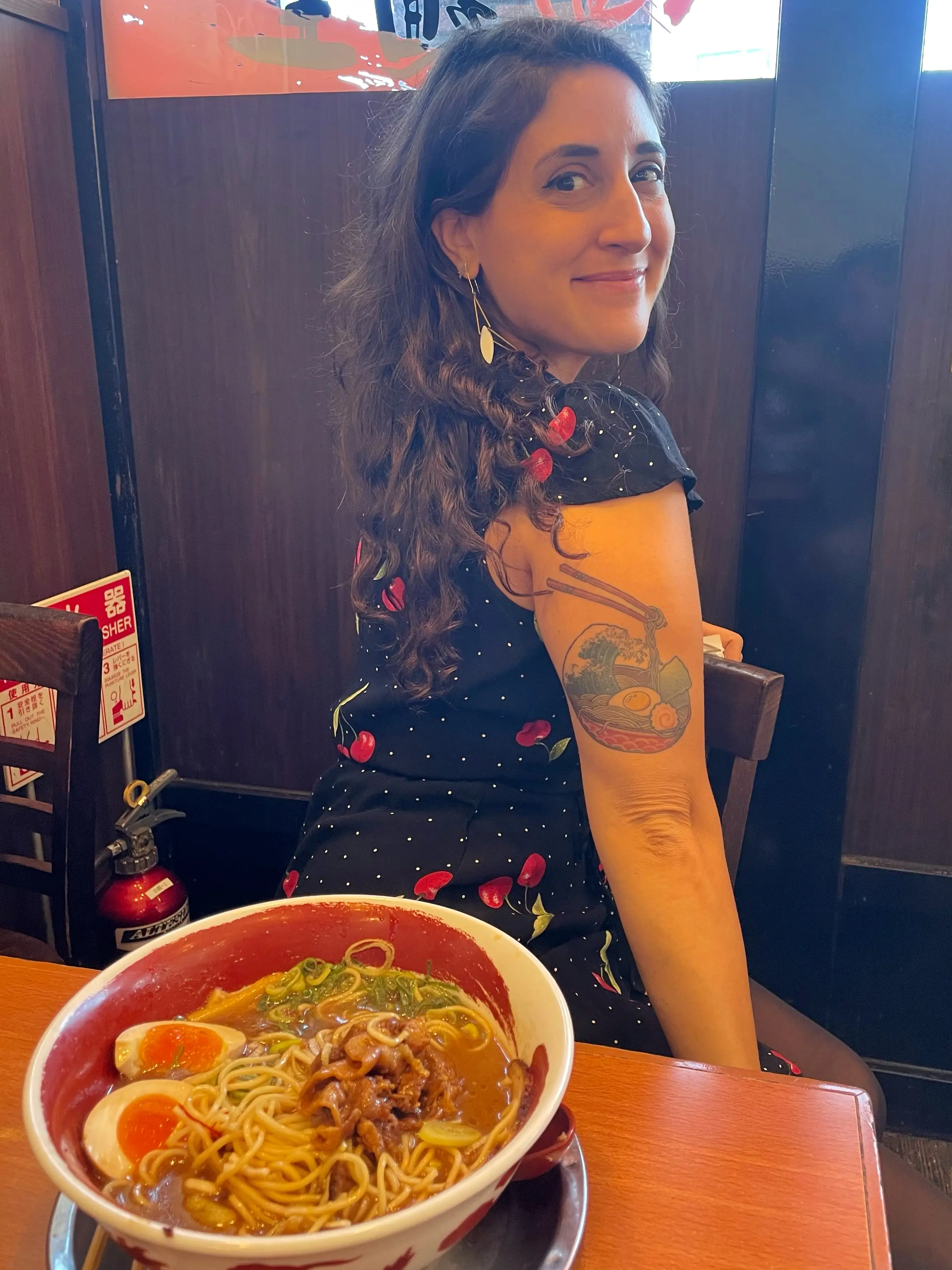 A woman is sitting at a table with a bowl of ramen, showing the ramen tattoo she has on her arm.