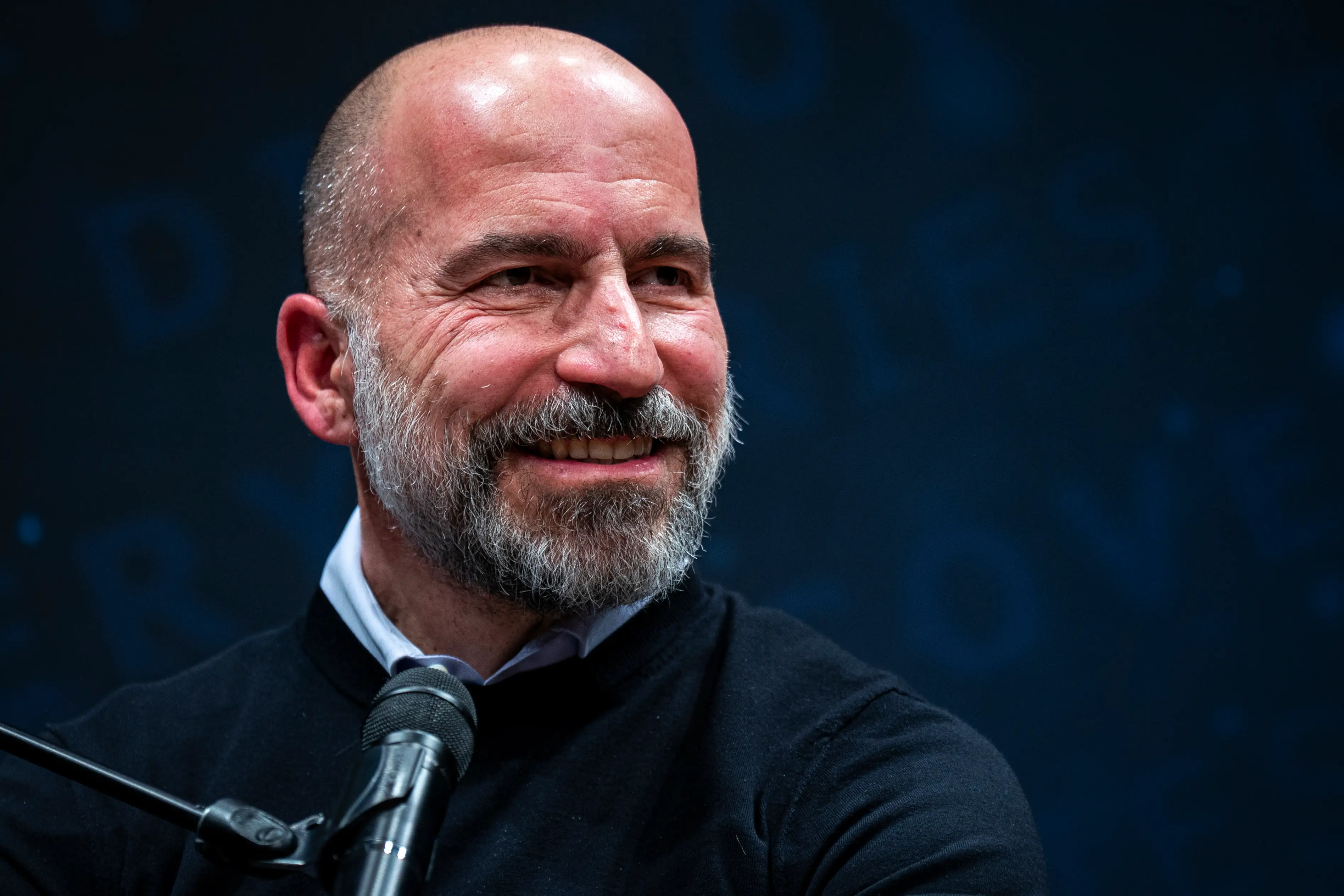 Uber CEO Dara Khosrowshahi smiles against a dark blue background while wearing a dark-colored sweater and sitting in front of a microphone.