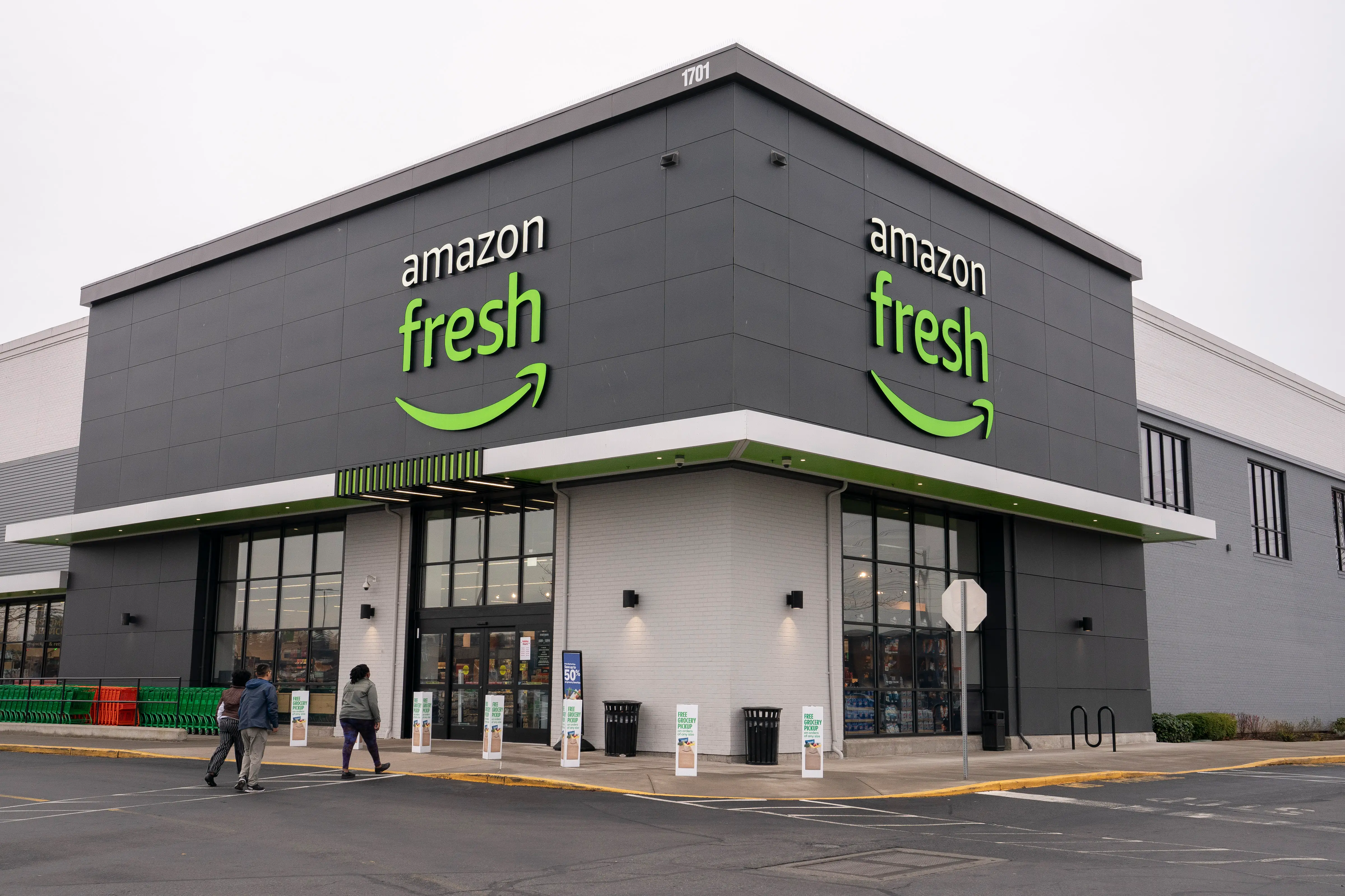 The entrance to an Amazon Fresh store, with green-and-white signs above doors leading into the store and a few customers walking in from the parking lot.