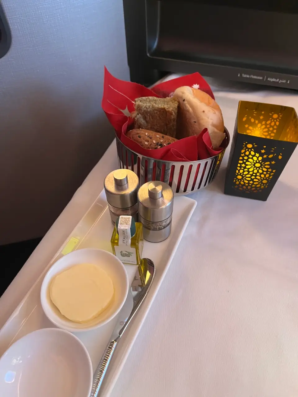 Bread basket next to small lantern, salt and pepper shakers, butter on tray table on Qatar Airways flight