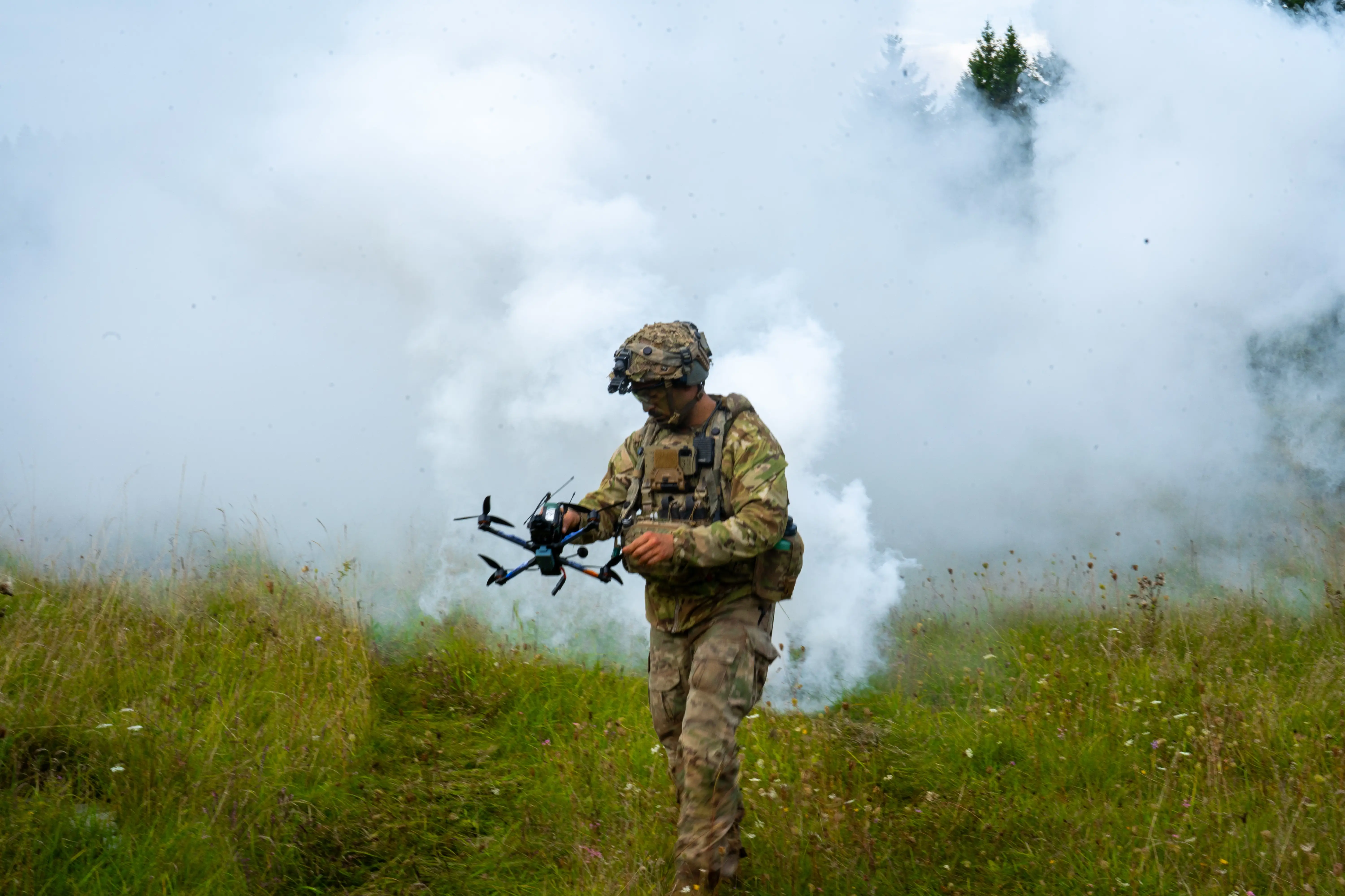 A soldier holds a quadcopter drone in a green field with walls of grey smoke behind him.
