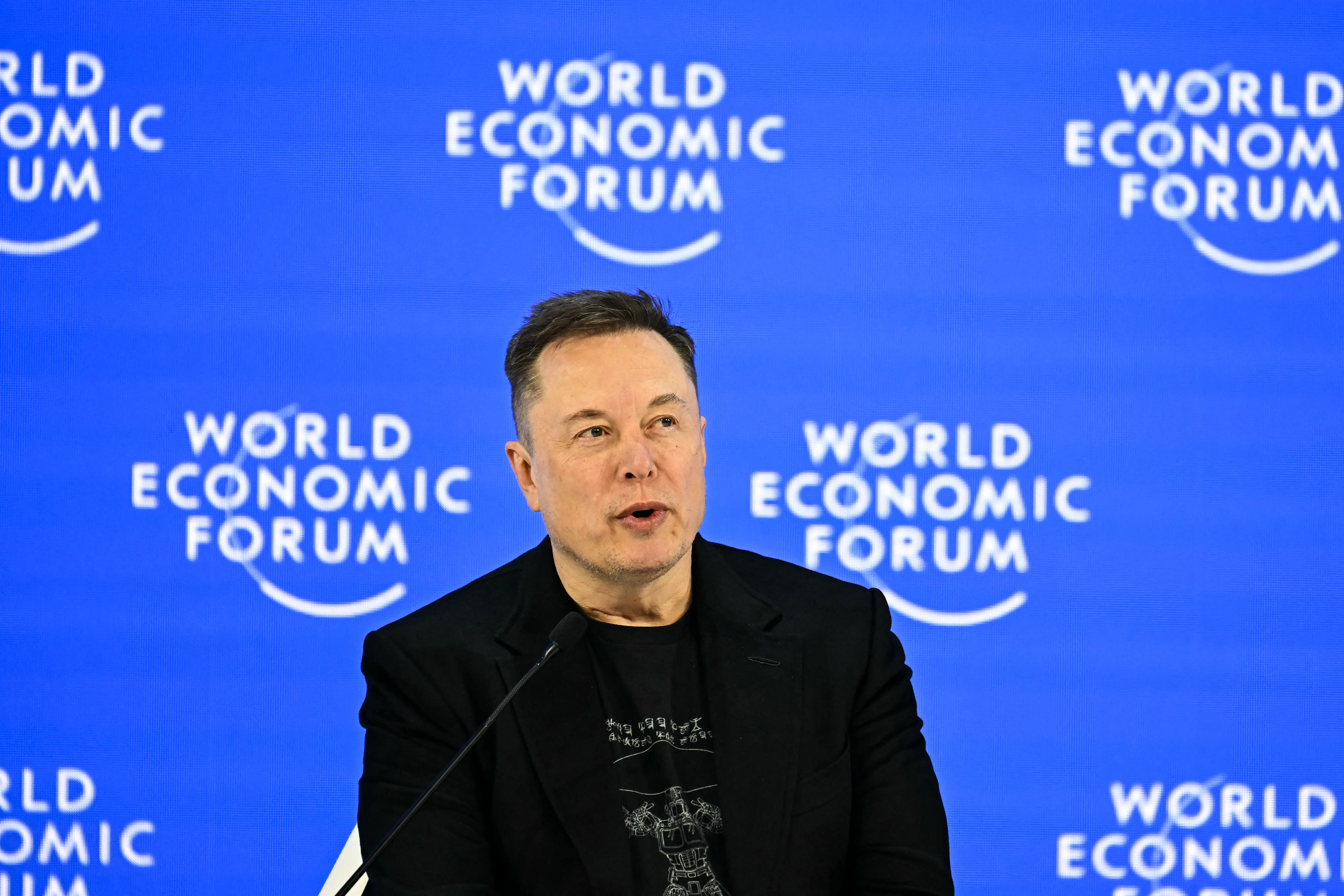 Elon Musk speaks on a panel at the World Economic Forum in Davos, Switzerland, against a blue backdrop.