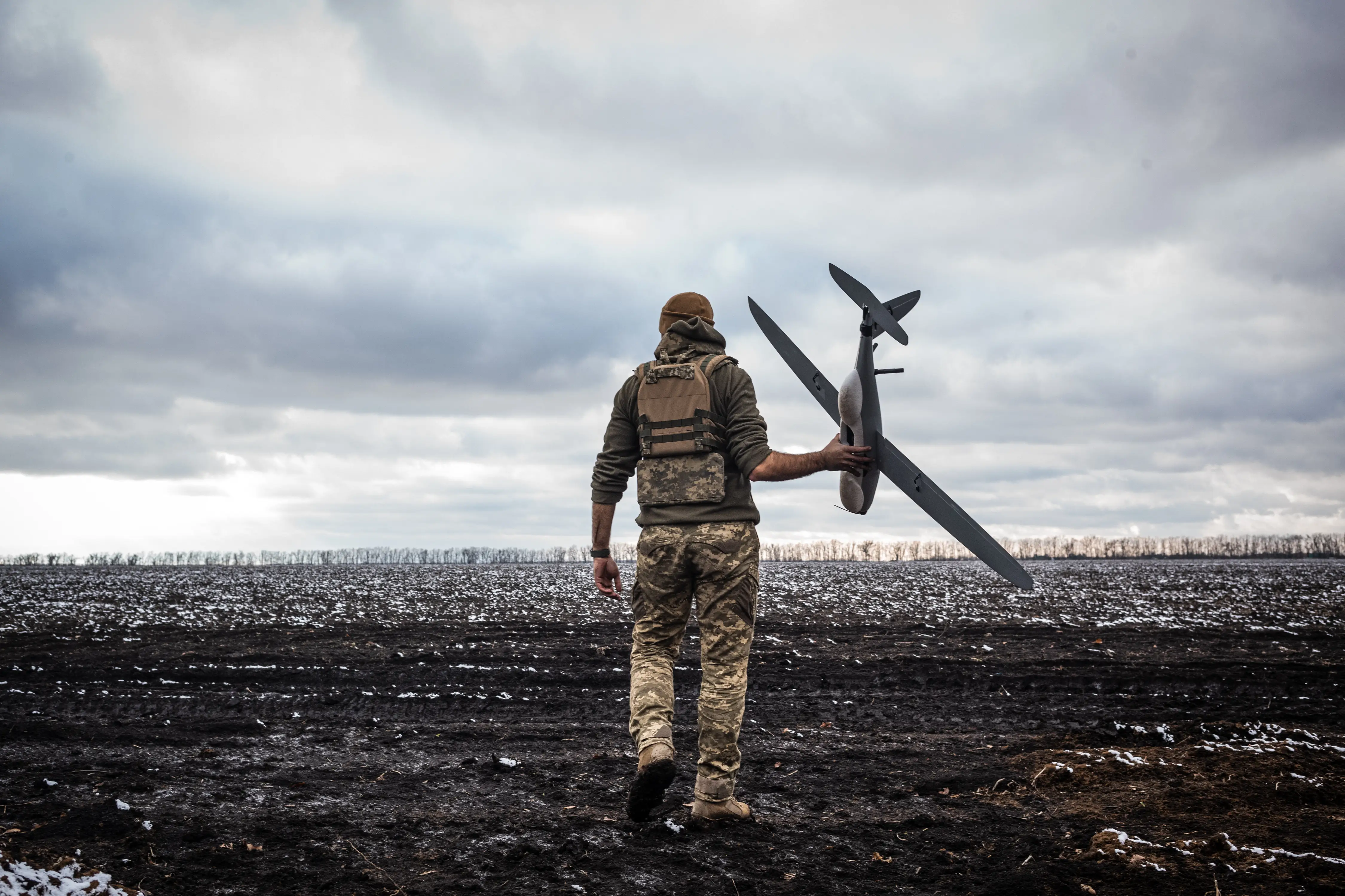 A man in camouflage gear with his back to the camera holds a drone, standing on muddy ground and beneath a grey sky