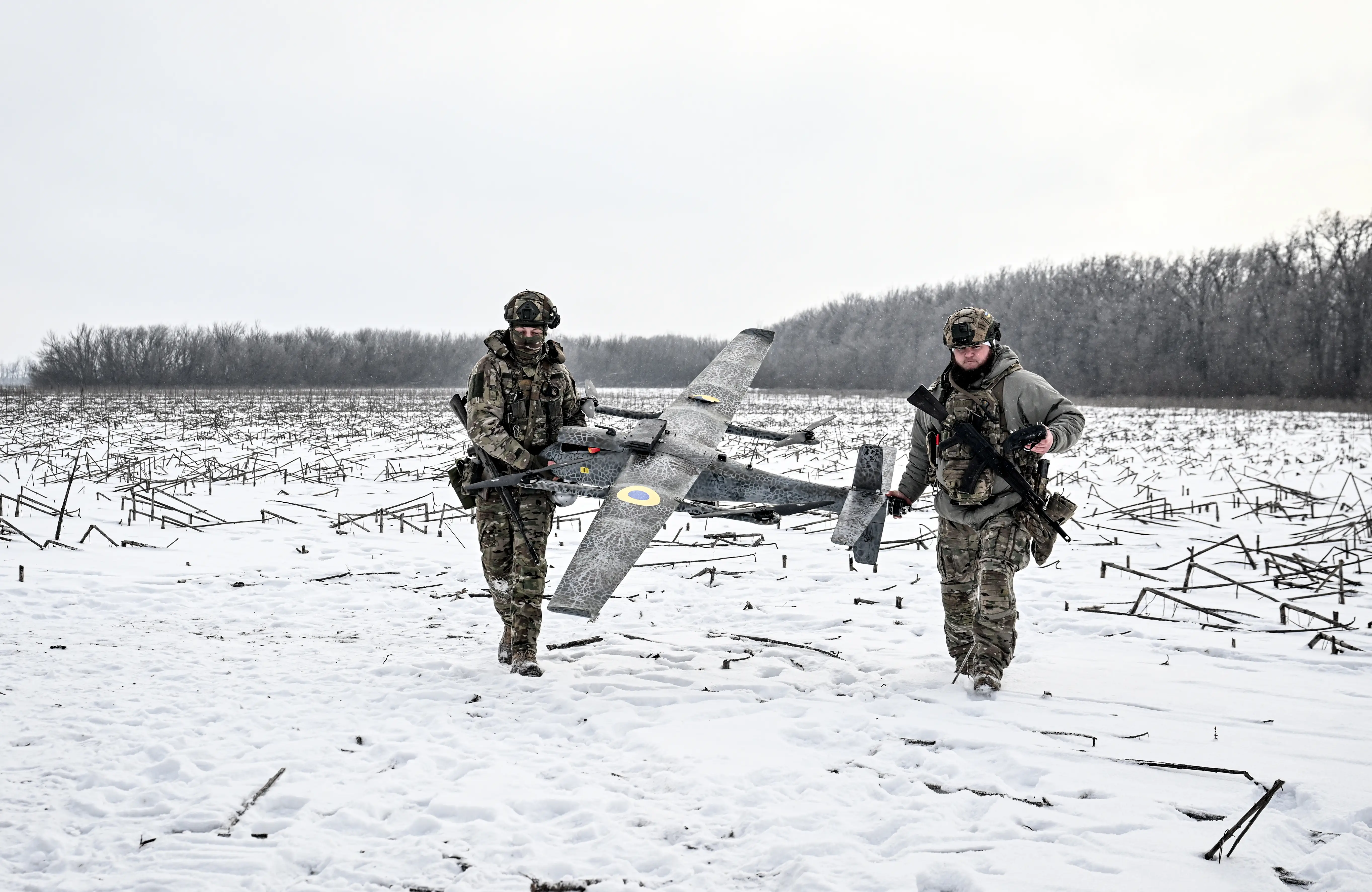 Two men in green camouflage gear walk across a snowy field holding a large grey drone, with a grey sky and trees behind them.