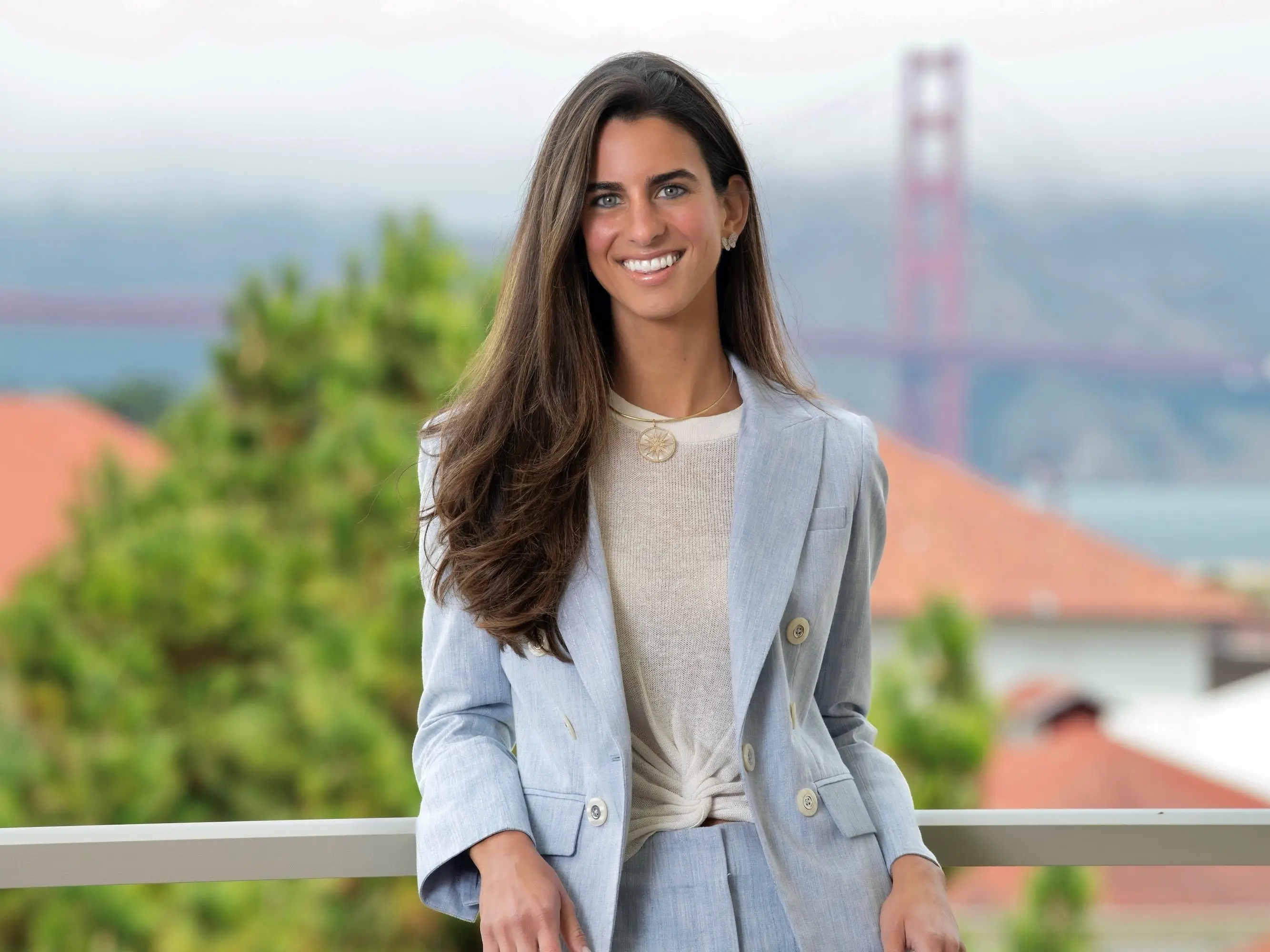 Smiling woman in a light blue suit leans on a railing outdoors with the Golden Gate Bridge blurred in the background.