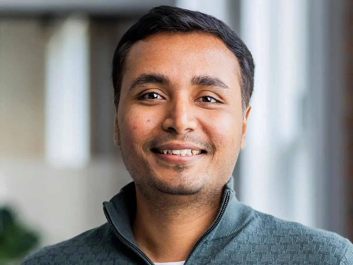 Smiling man with short dark hair wears a gray quarter-zip sweater in a close-up portrait with a softly blurred indoor background.
