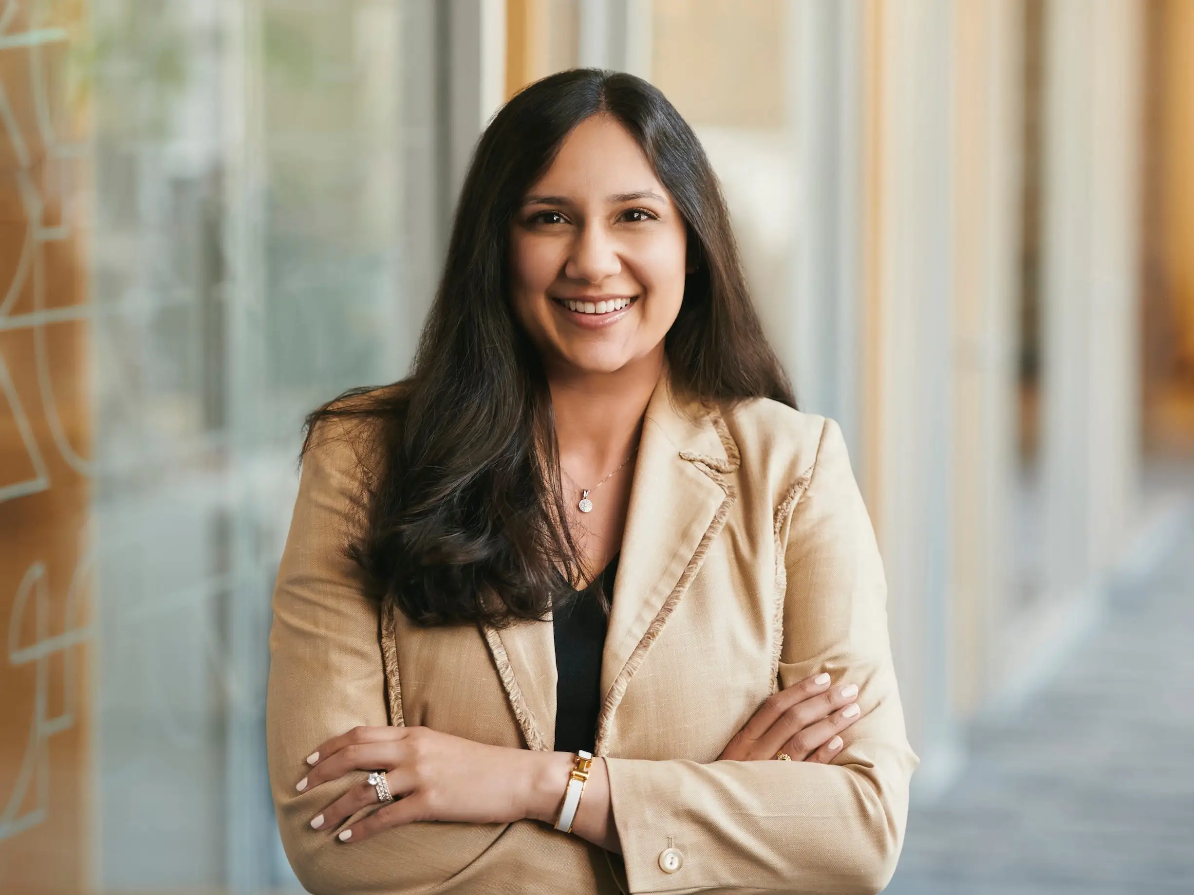 Smiling woman with long dark hair wears a tan blazer and stands with her arms crossed in a bright office hallway with glass walls.