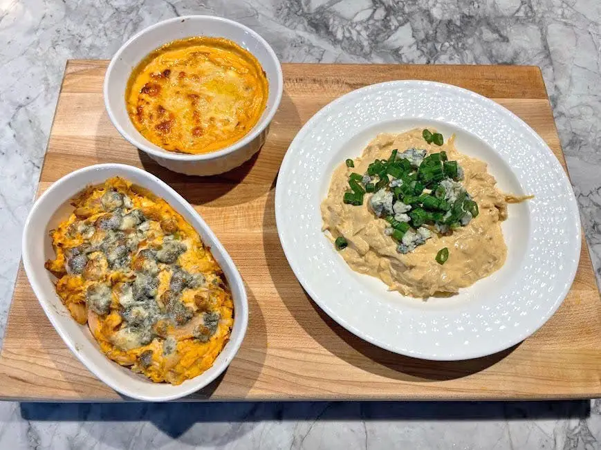 Straight-on shot of a cutting board with three bowls and plates of Buffalo-chicken dips. One bowl has a dip with crumbled blue cheese on top, one bowl has a dip with melted yellow cheese, and one dip is topped with crumbled blue cheese and scallions.