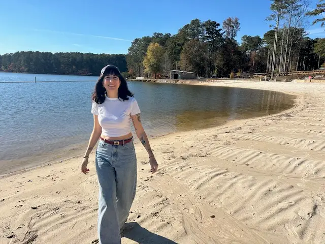 Woman in white shirt and jeans walking away from water on sand, smiling