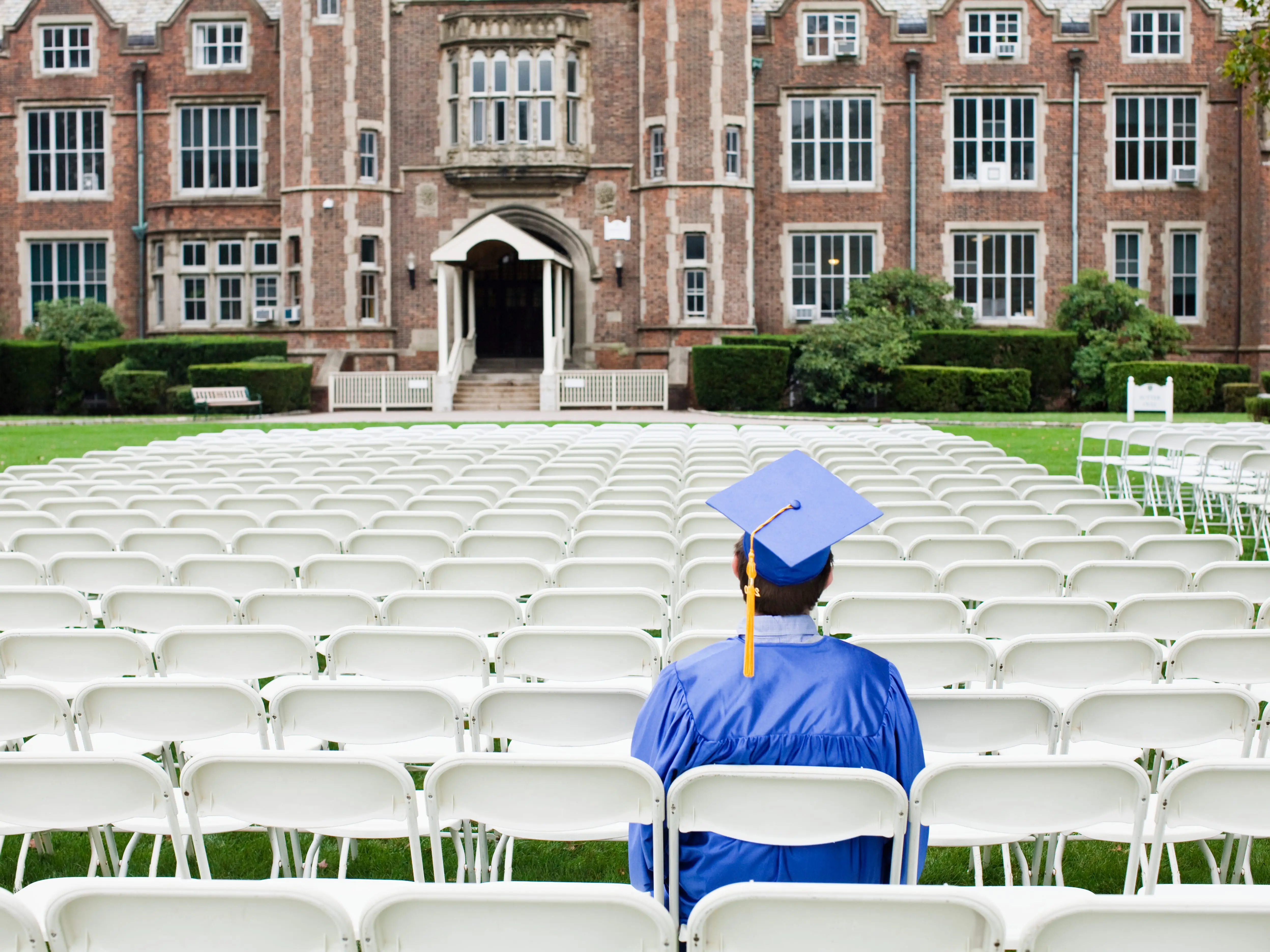 A graduate wearing blue is sitting in one of many chairs outside, facing a building