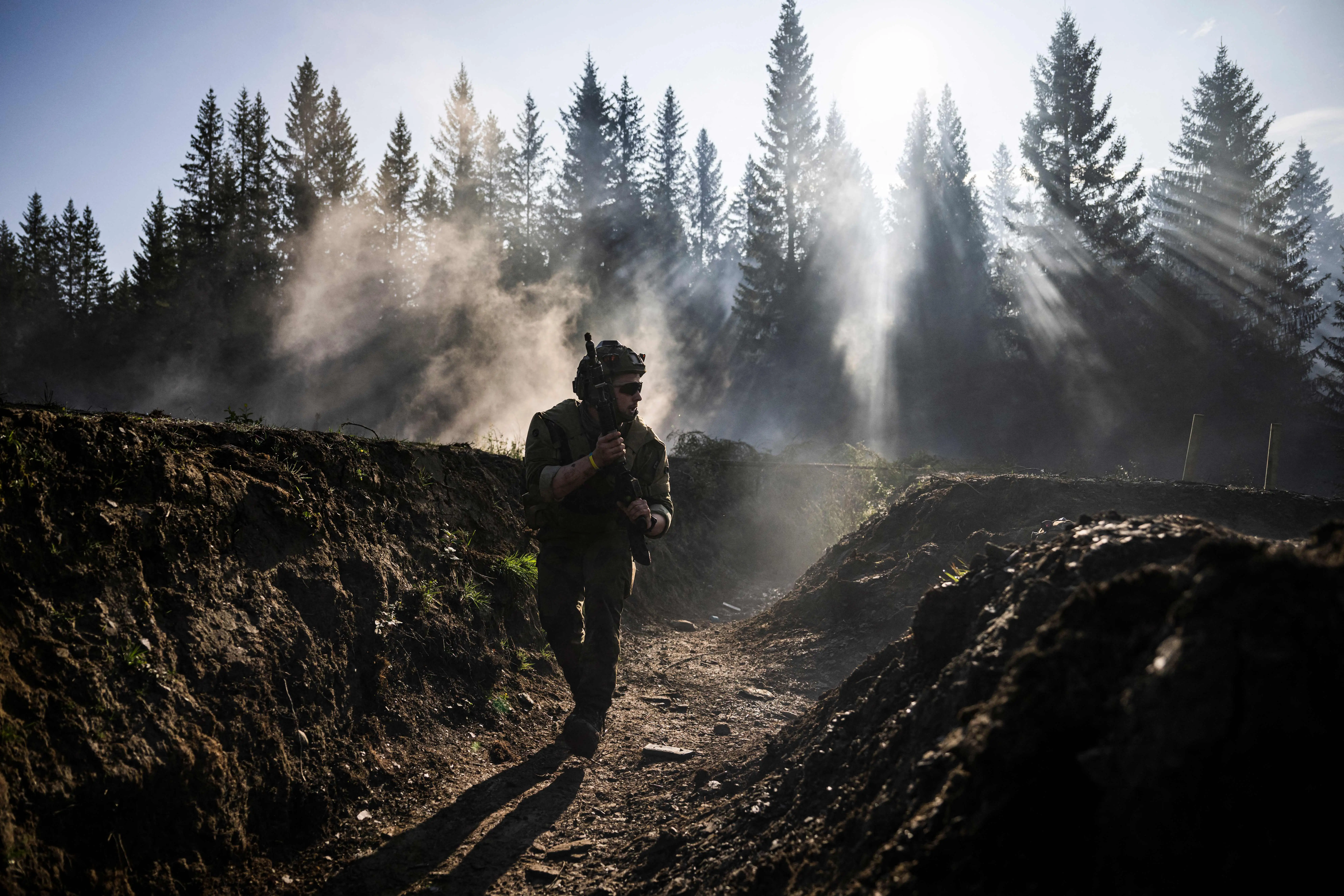 A figure holding a firearm moves through a trench with a foggy blue sky and trees behind
