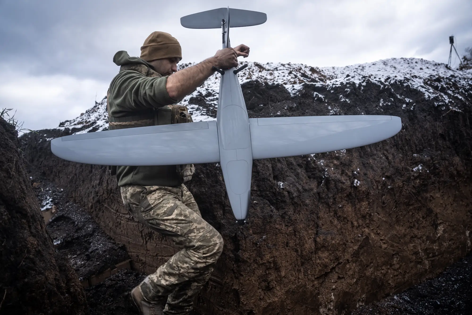 A man in camouflage clothing and a beanie moves through a snowy trench holding a large grey drone by its tail