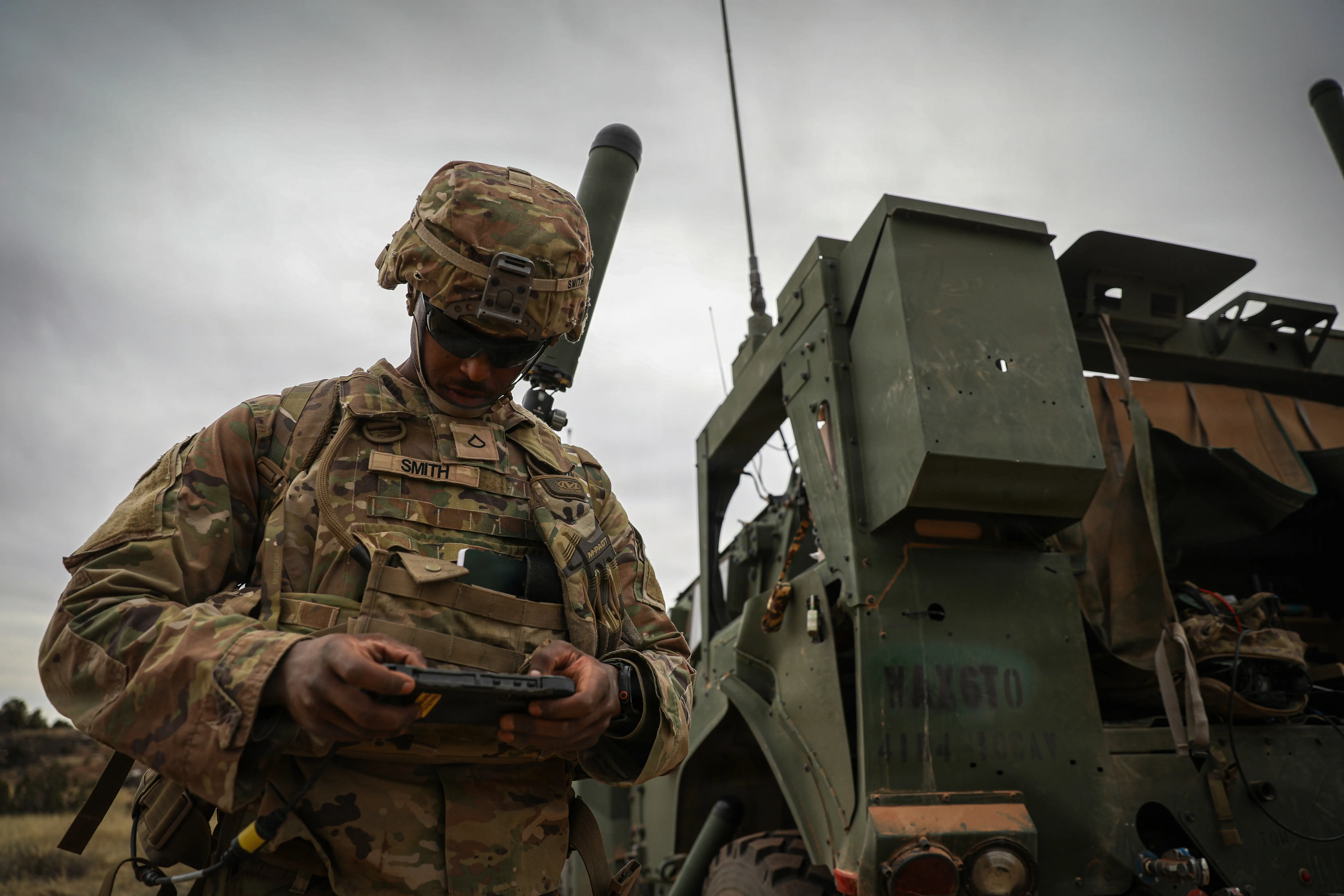 A soldier looks down at a tablet while standing next to a vehicle.