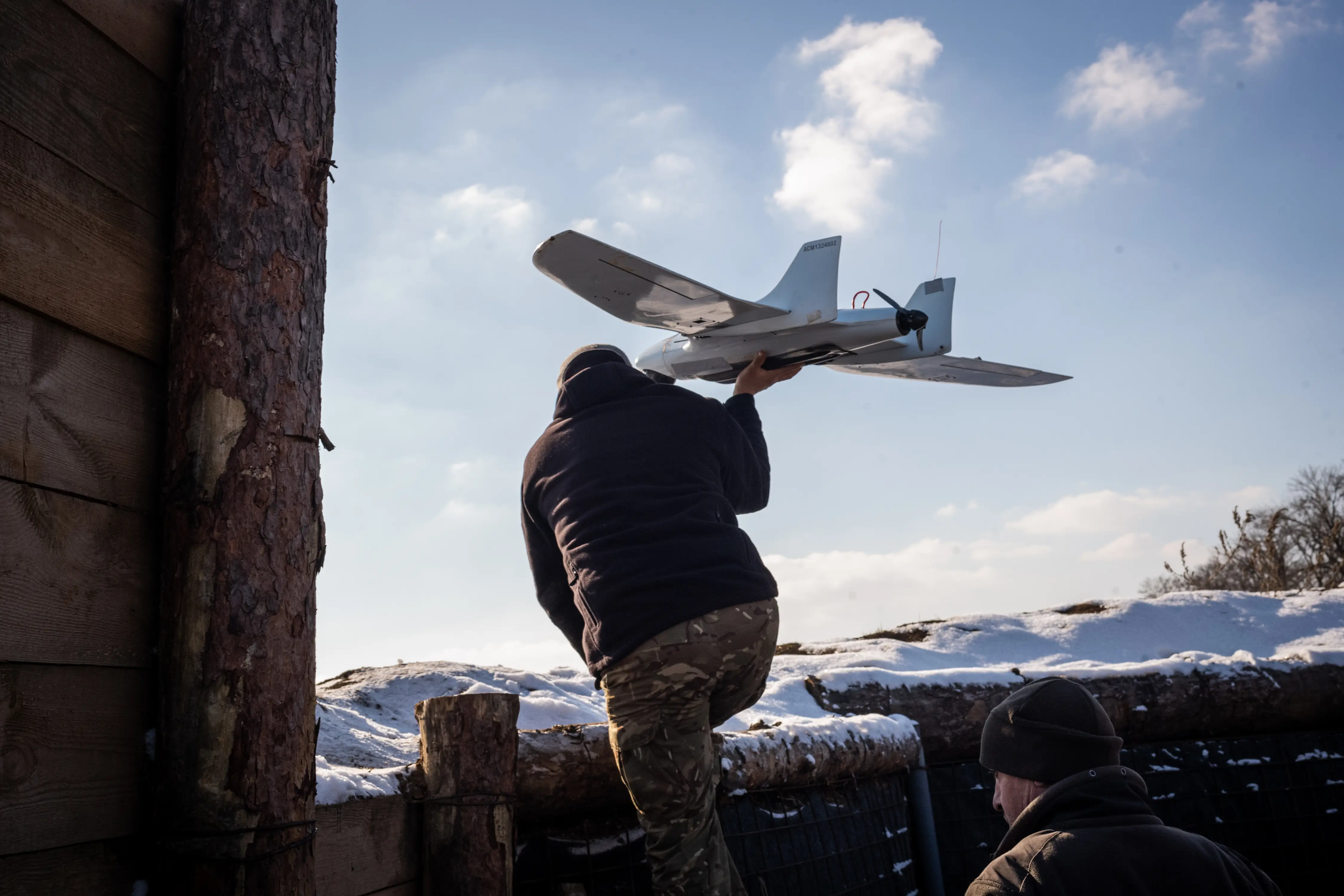 The back of a figure holding a grey drone climbs out of a wooden trench-like structure with snow and blue sky in front of him
