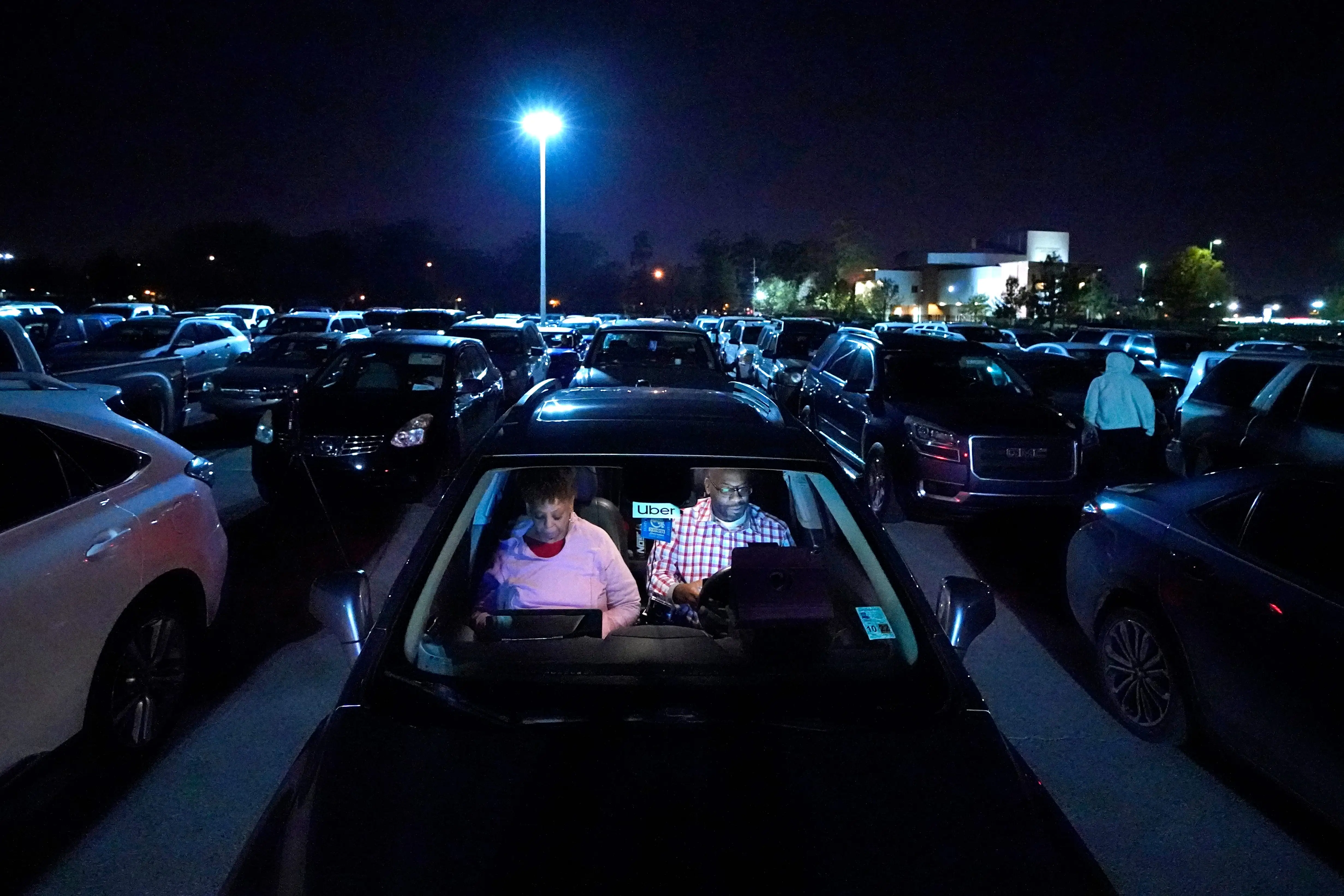 Two people sit in a car with an Uber sign in the windshield while parked around other cars at night with a streetlight in the background