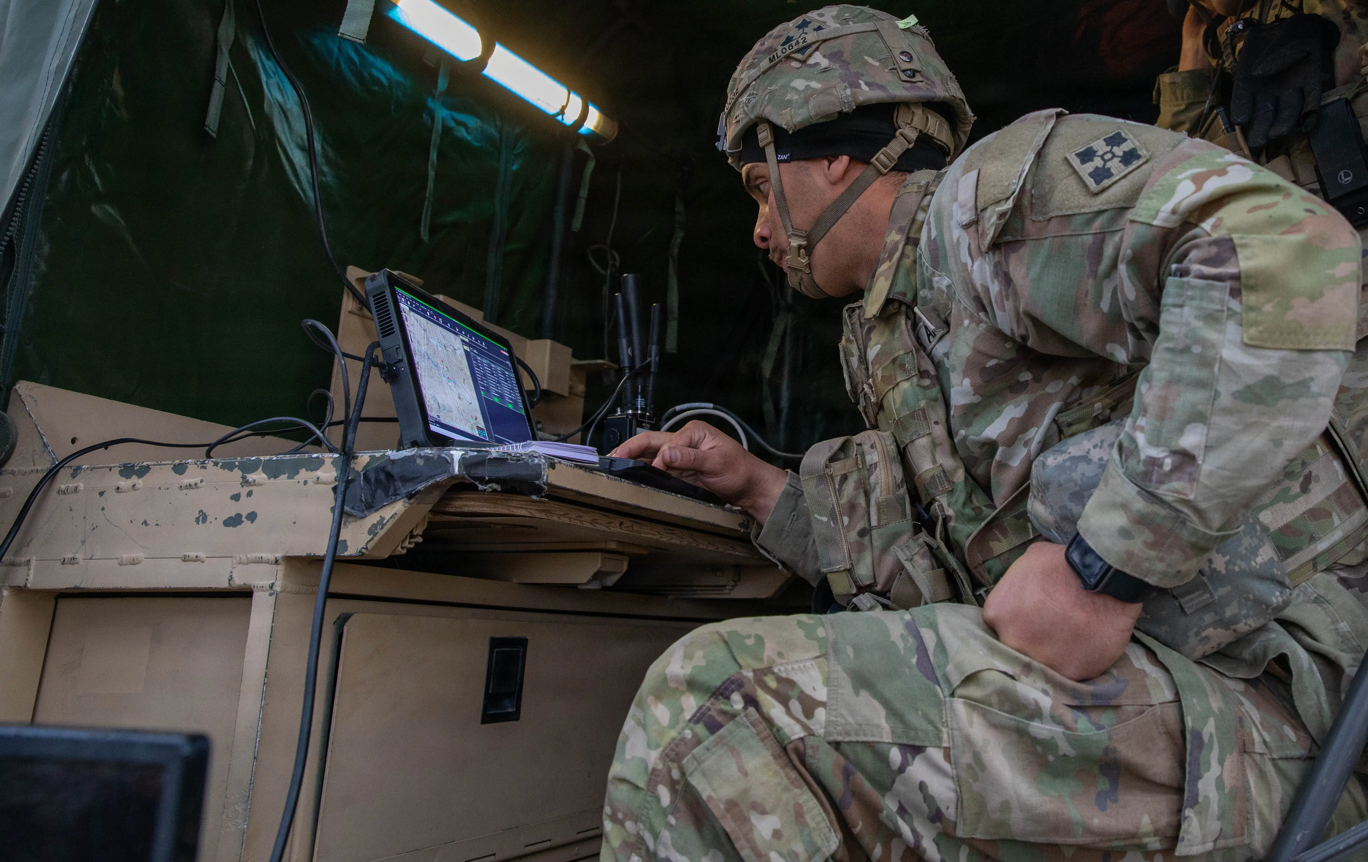 A soldier sits at a desk and looks at a computer inside a tent.