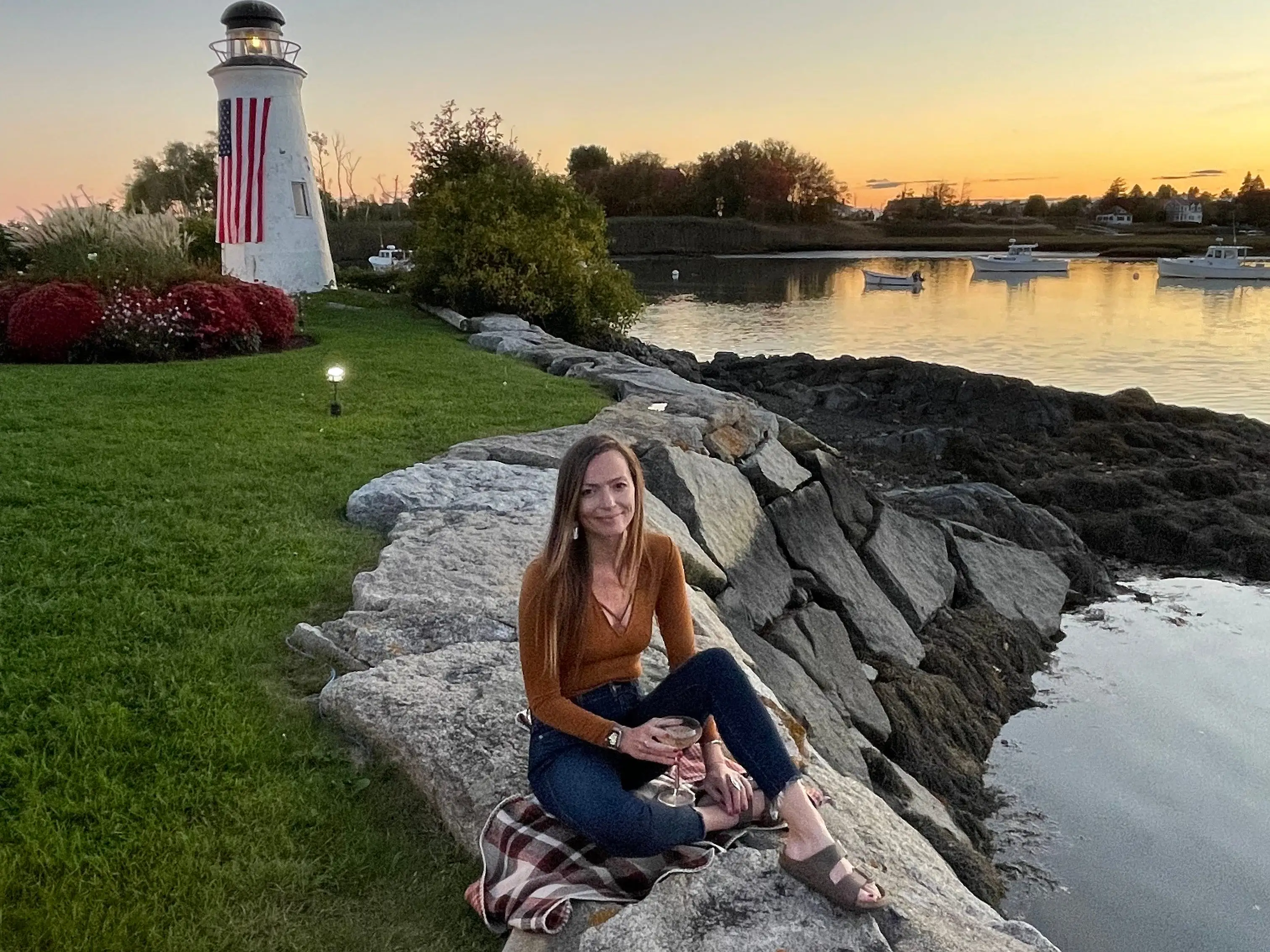 Emily sits on rocks near the water in Kennebunkport, Maine, with a lighthouse in the background.