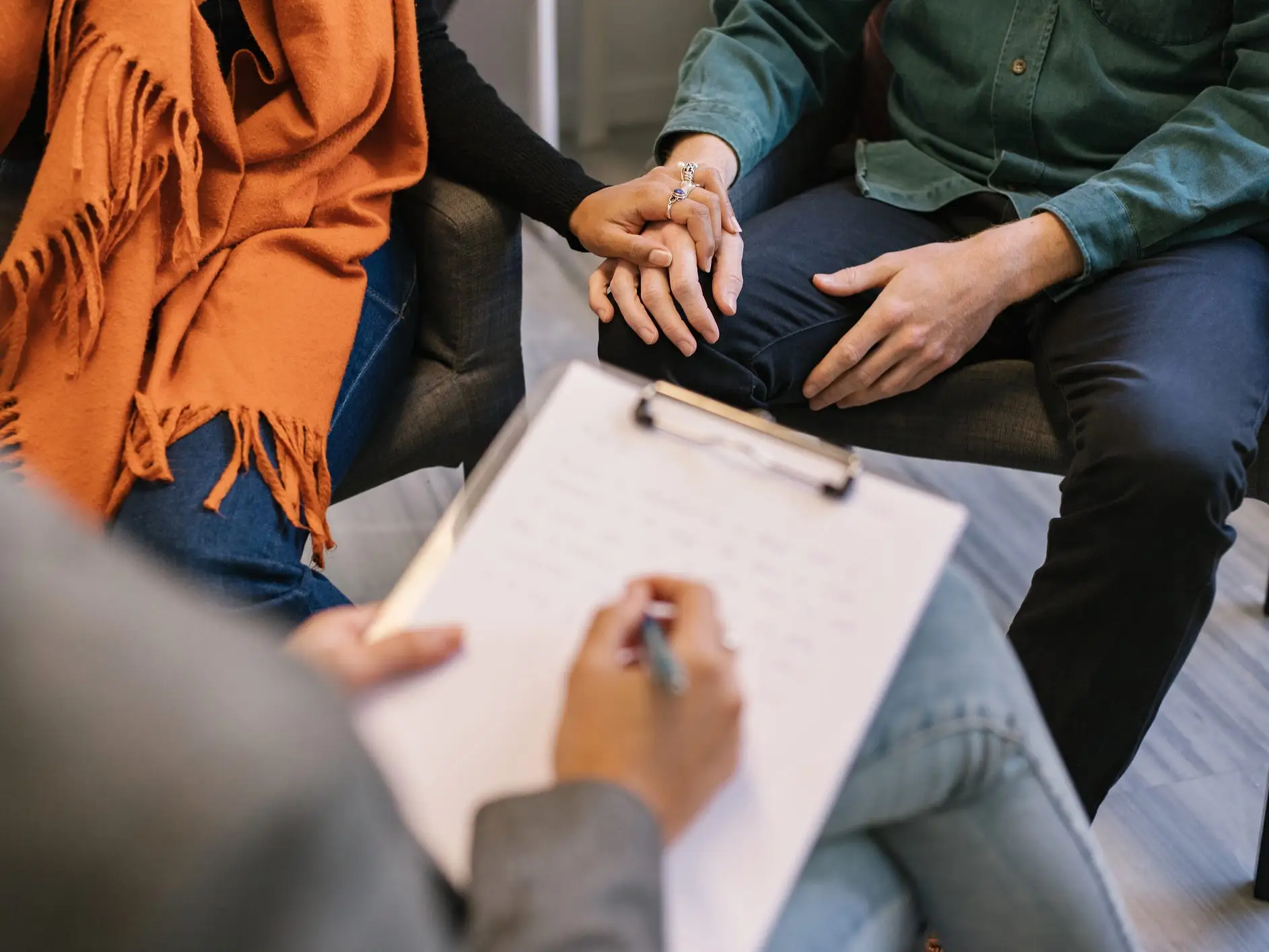 Couple sharing marriage problems and holding hands while therapist taking notes in notepad on psychology session