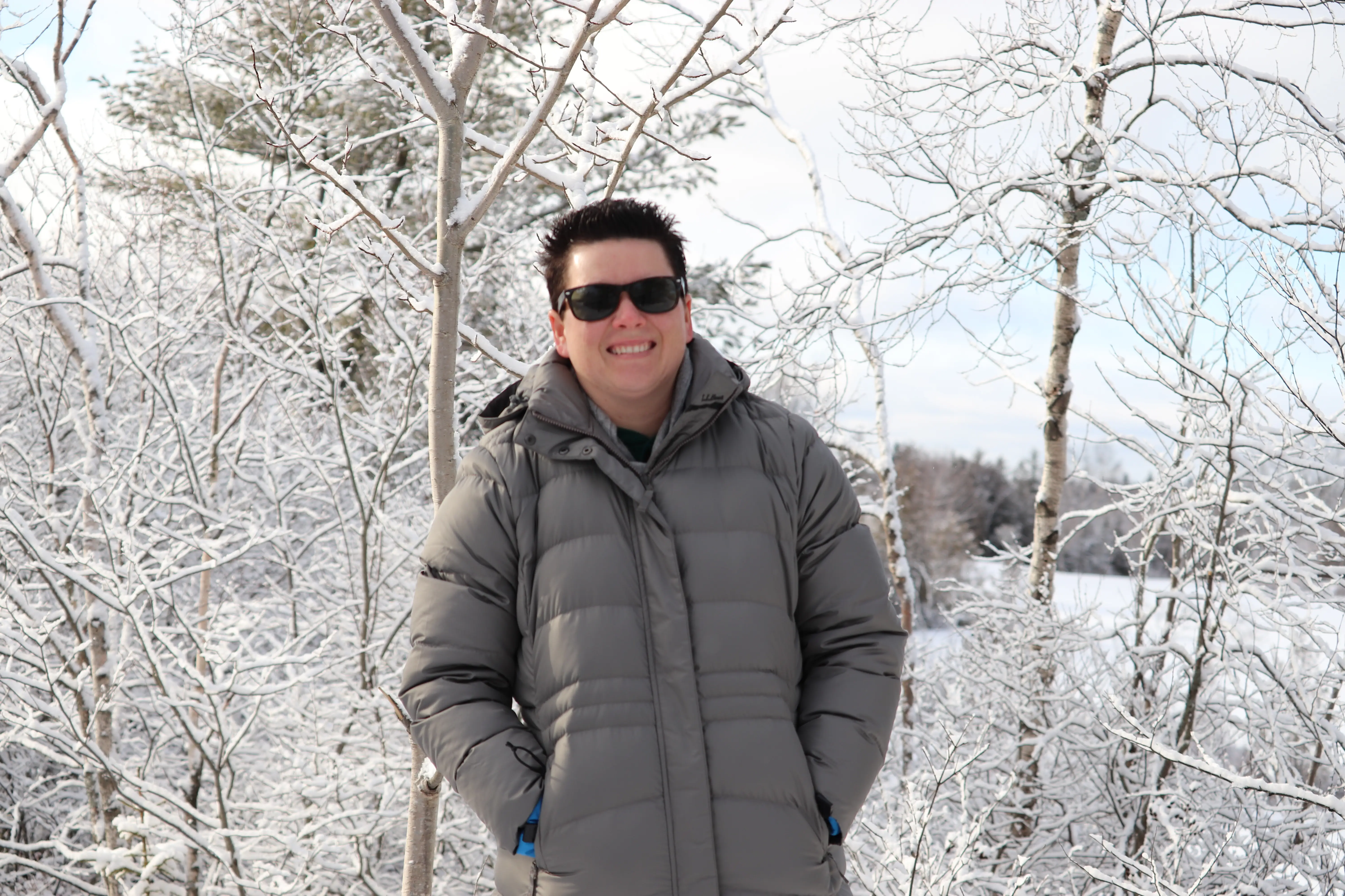 Woman in jacket and sunglasses smiling in front of snow-covered trees
