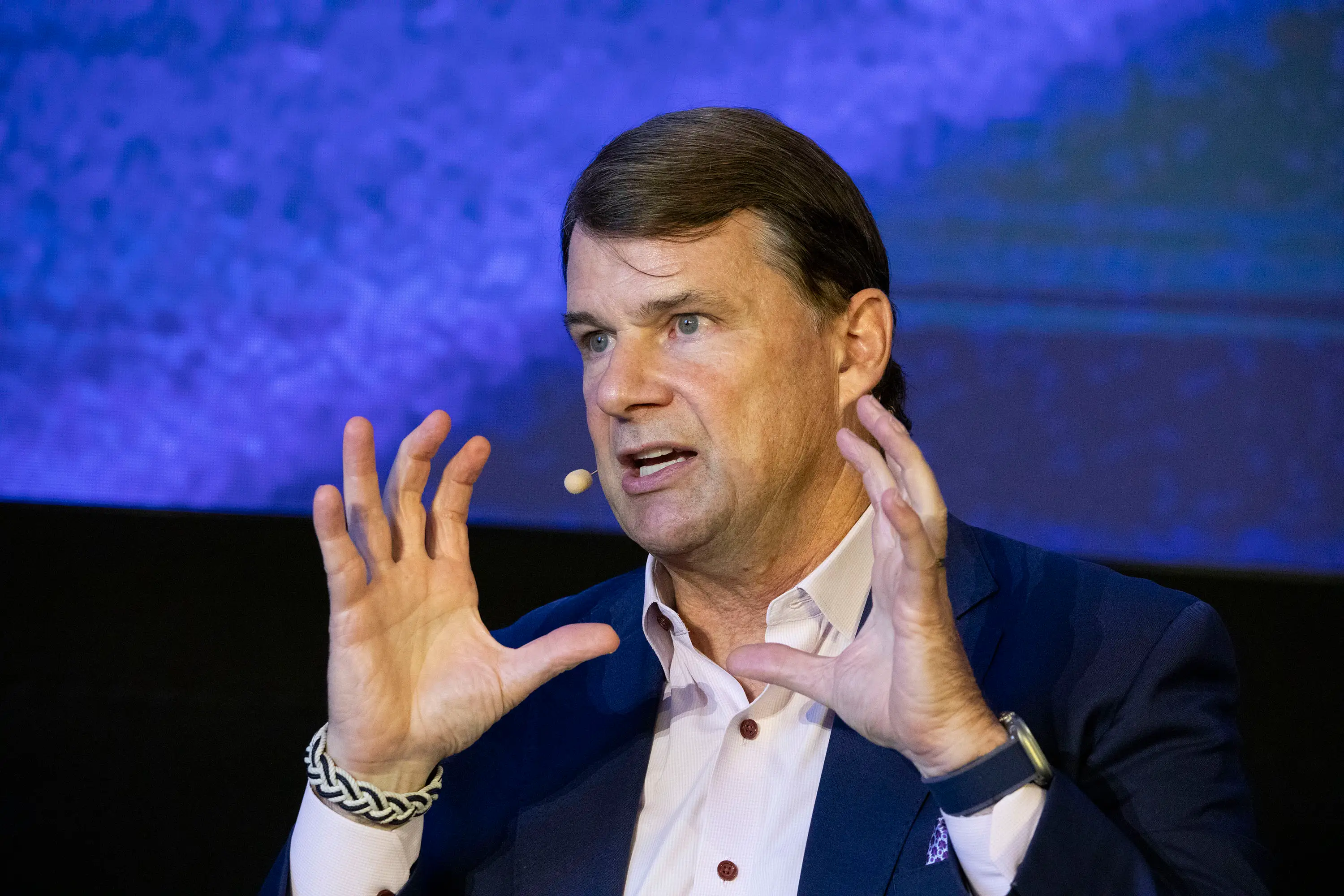 Jim Farley is speaking on stage in front of a blue wall. He is wearing a blue suit and a white shirt.