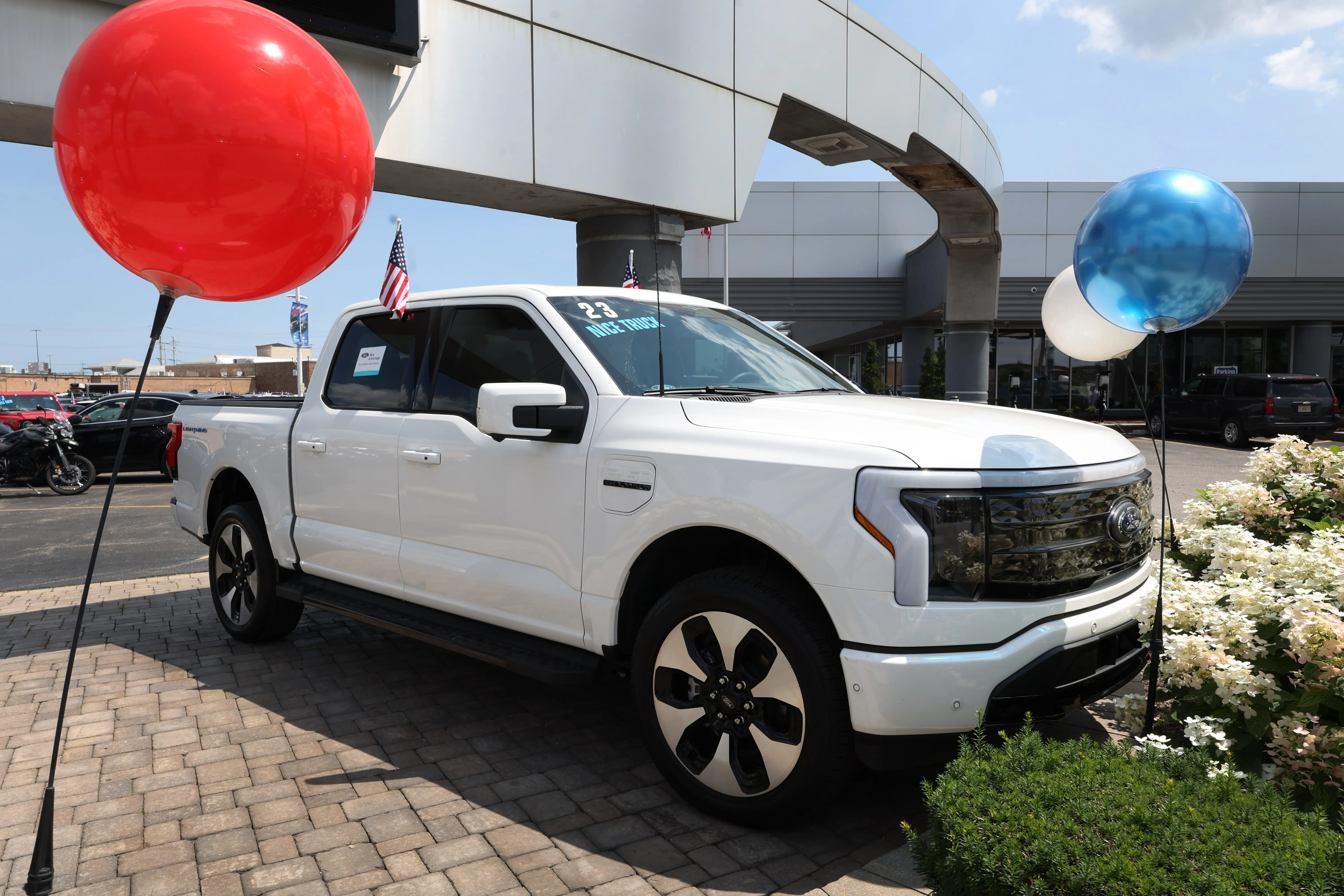 A white Ford F-150 Lightning pickup parked outside a dealership. The truck is surrounded by balloons and flowers and has an American flag flying from the second-row window.