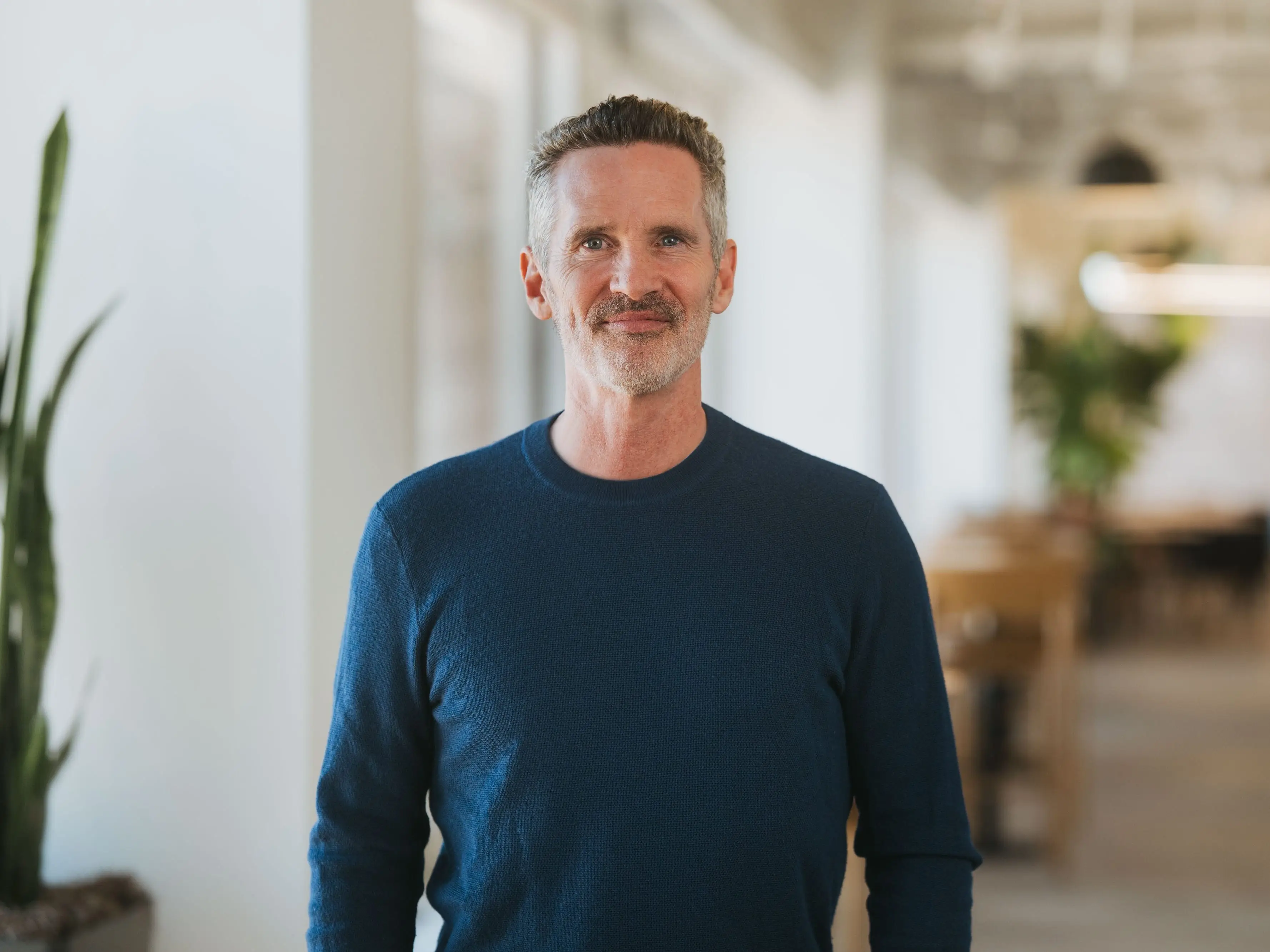 A man with short gray hair smiles at the camera while standing in a bright, modern office.