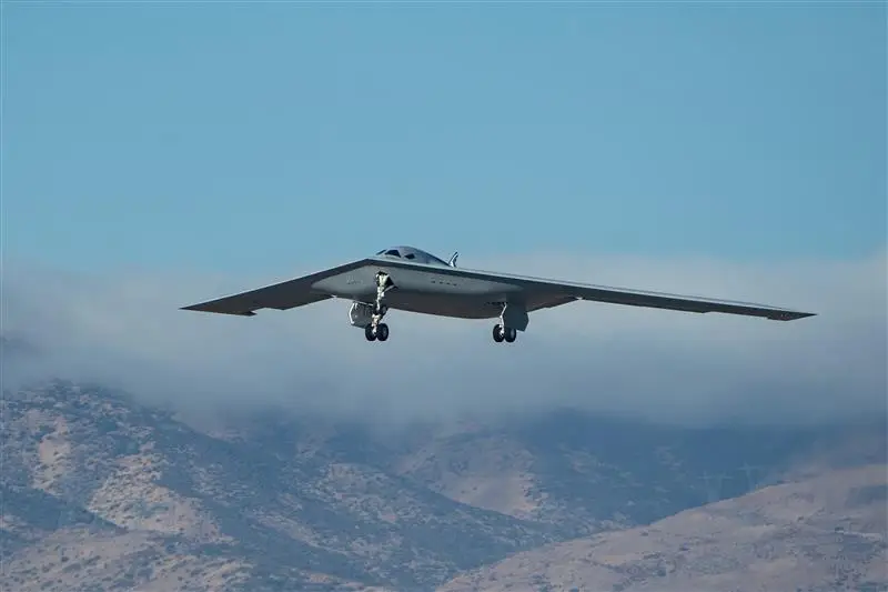 A B-21 Raider bomber flies above a mountainous terrain with clouds and a clear blue sky in the background.